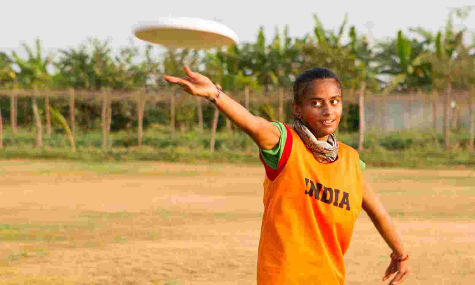 印度隊最佳 Ultimate Frisbee 背景 壁紙 — 一位身穿橙色印度球衣的年輕女性在塵土飛揚的場地上擲出白色飛盤，背景有棕櫚樹。