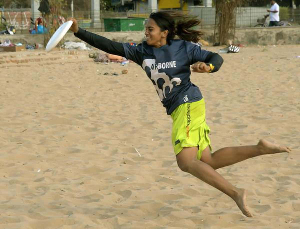 印度飛盤運動員 Smithi Manickam 最佳 Ultimate Frisbee 背景 壁紙 — 一個快樂的女人在沙灘上跳起，拋出白色飛盤，動作充滿動感，穿著鮮綠色短褲。