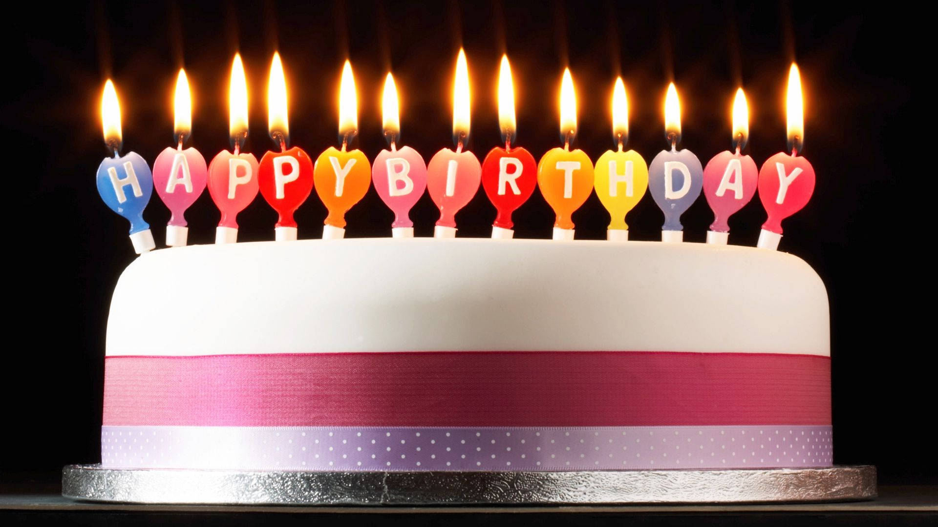 印有「生日快樂」蠟燭的生日蛋糕 壁紙 — 一個白色生日蛋糕，上面有彩色心形蠟燭拼出「生日快樂」，背景為黑色。