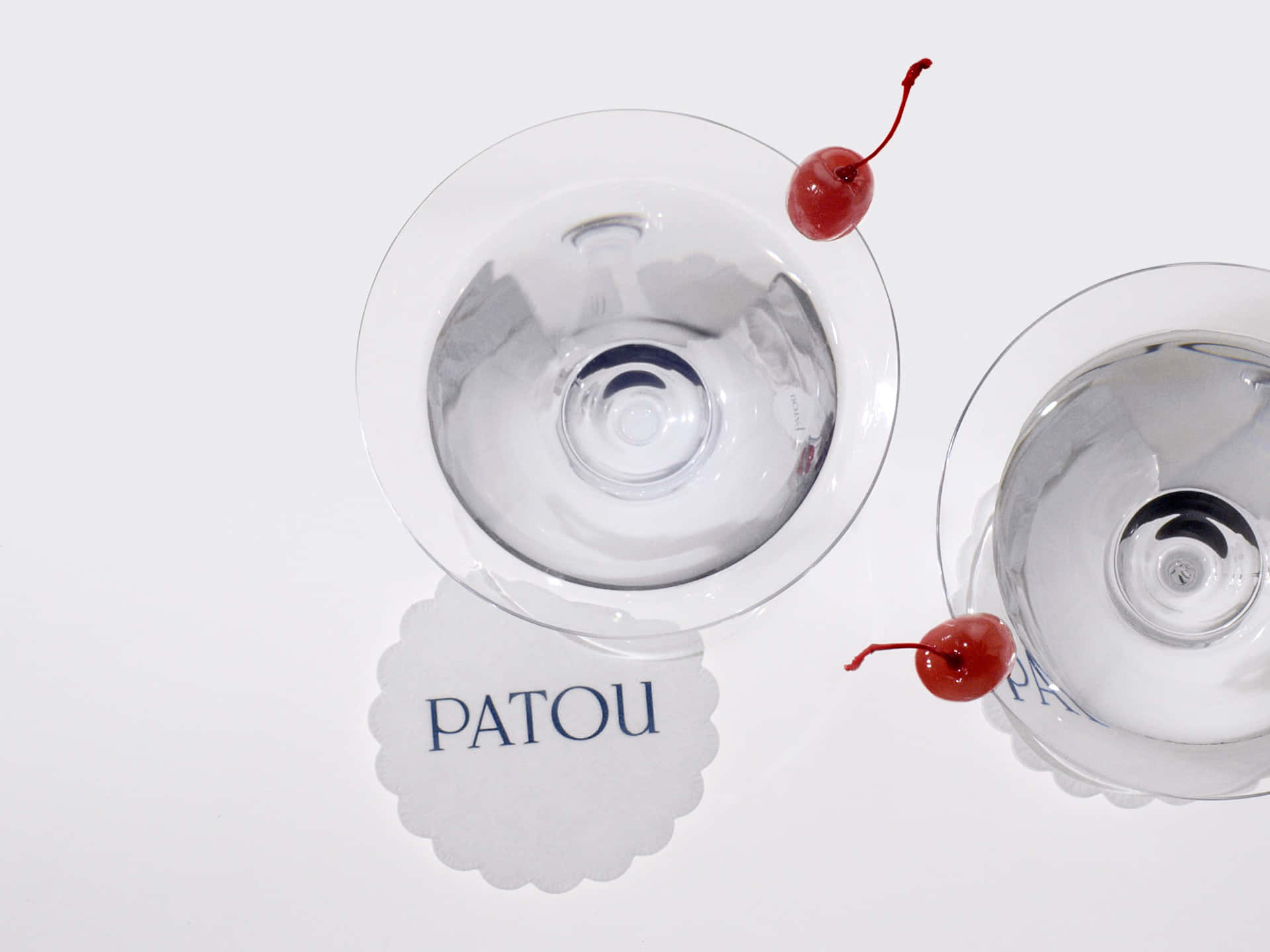 印有Patou標誌的蕾絲墊上的雞尾酒杯平鋪圖 壁紙 — 兩個空馬提尼杯，杯中有帶梗櫻桃，放在印有品牌標誌的杯墊上，背景為純白色。