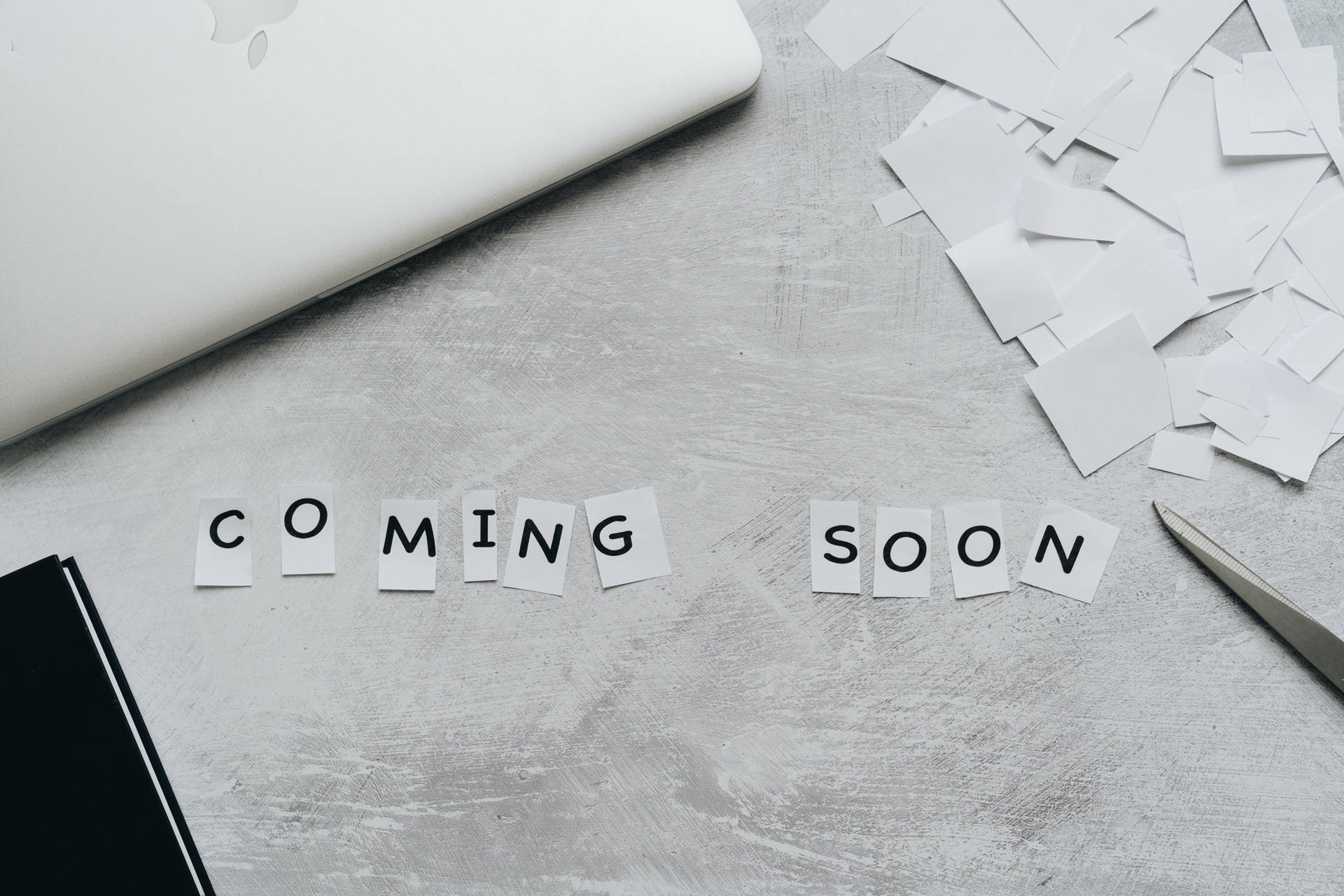 灰色桌面上，手提電腦和剪刀旁，用紙字母拼出coming soon。