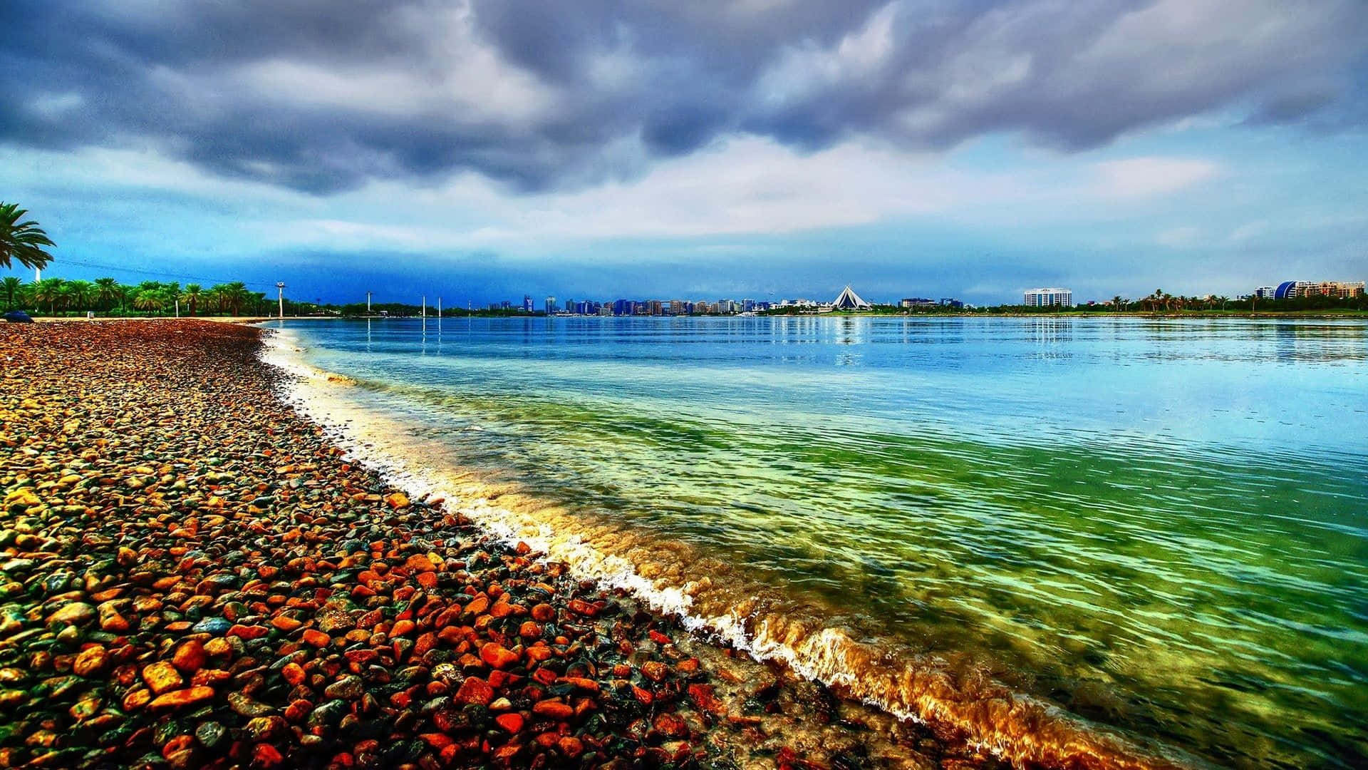 卵石海岸海灘主題背景 壁紙 — 充滿活力的海岸線，色彩繽紛的鵝卵石與碧綠的海水相遇，在戲劇性的多雲天空下，遠處的城市天際線。