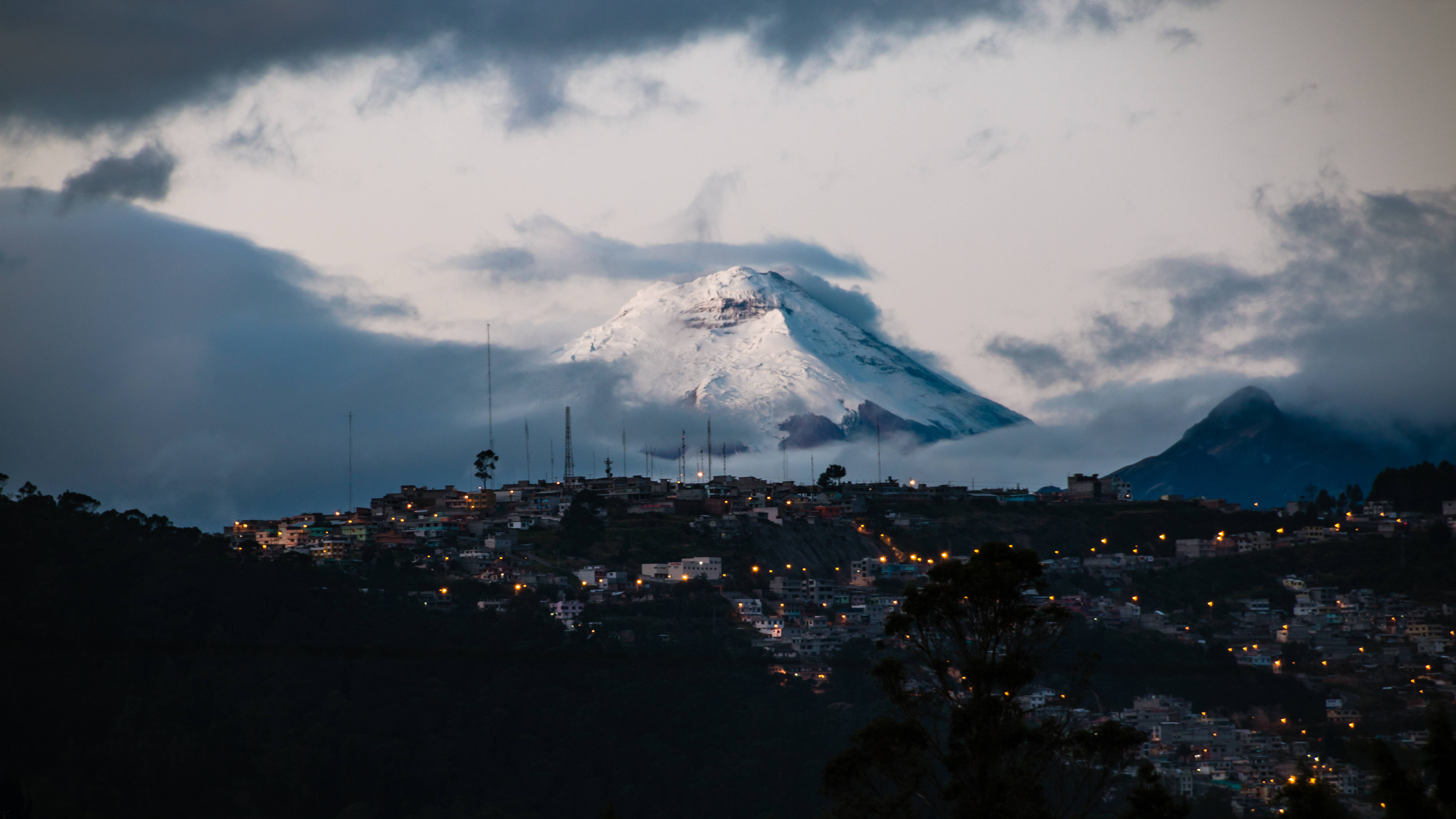 厄瓜多爾科托帕克火山 壁紙 — 一座積雪的山峰聳立於山丘小鎮之上，小鎮在多雲的暮色天空下閃爍著溫暖的燈光。