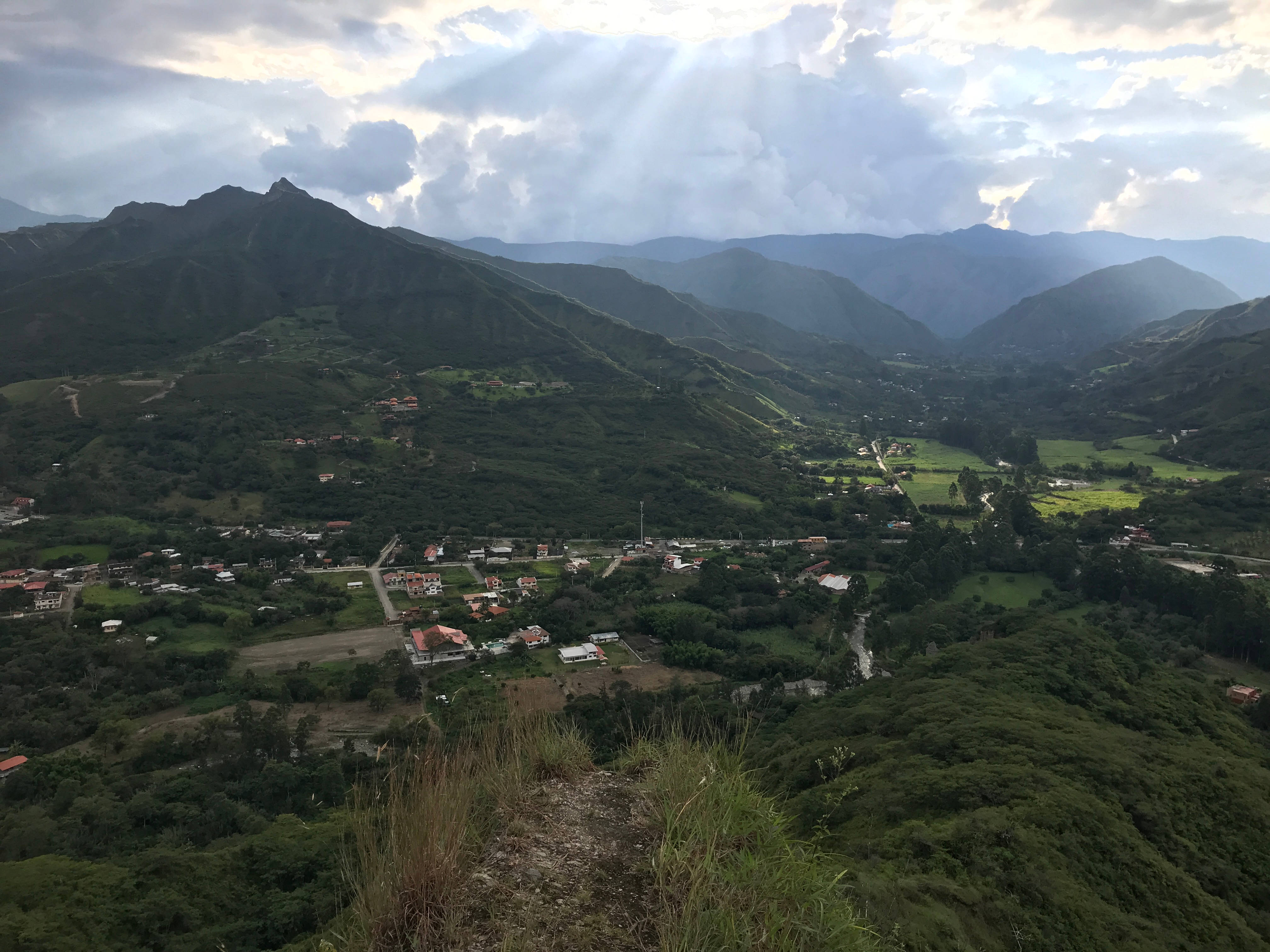 厄瓜多爾維拉卡班巴村莊 壁紙 — 陽光穿過雲層，照亮了一個鬱鬱蔥蔥的綠色山谷，山谷中有一個小村莊坐落在連綿起伏的山巒之間。