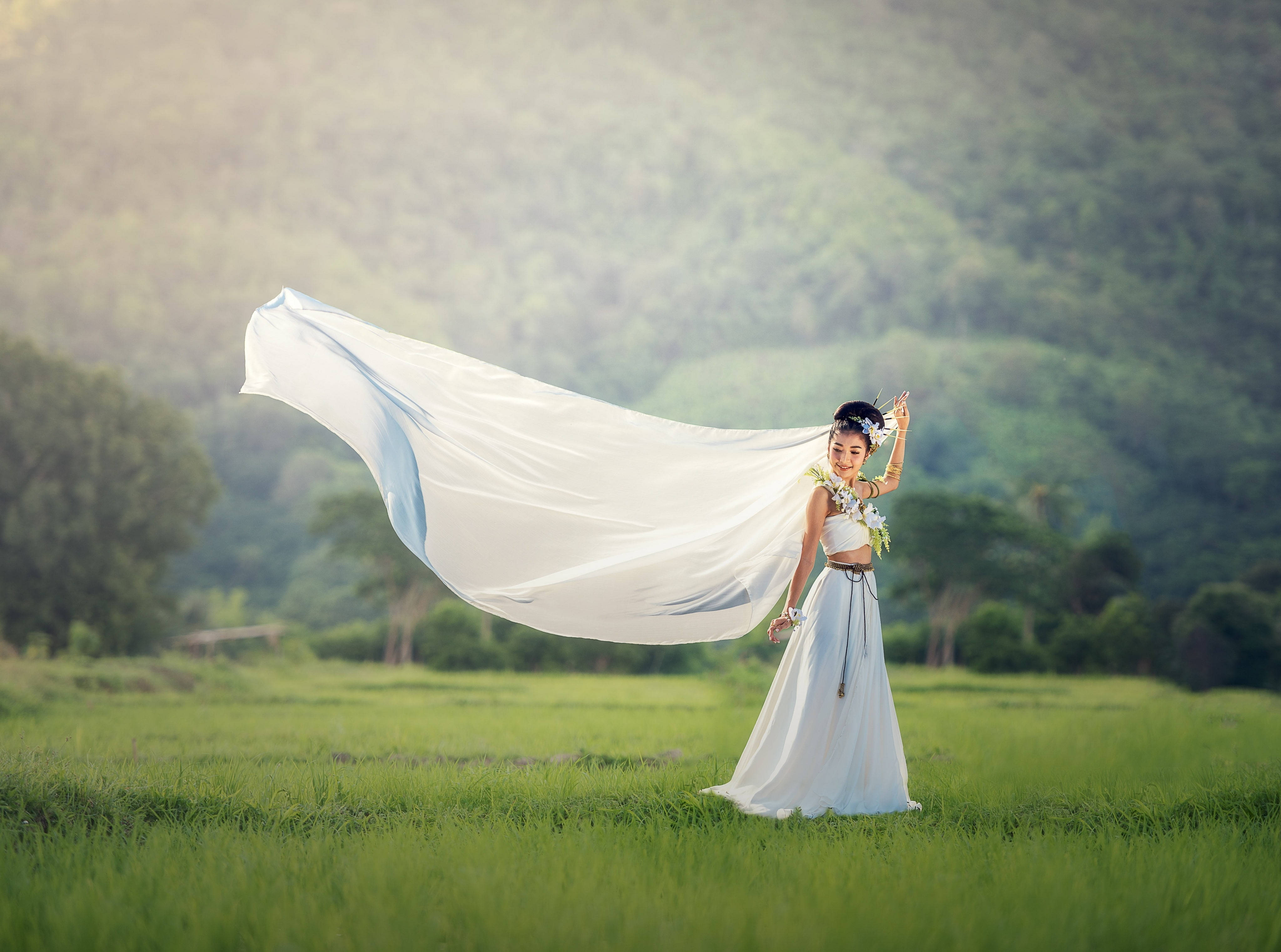 原住民婚紗 壁紙 — 一位身穿飄逸白色連衣裙的女性，站在陽光明媚的綠色田野中，身後是山脈，她輕薄的布料隨風飄揚。