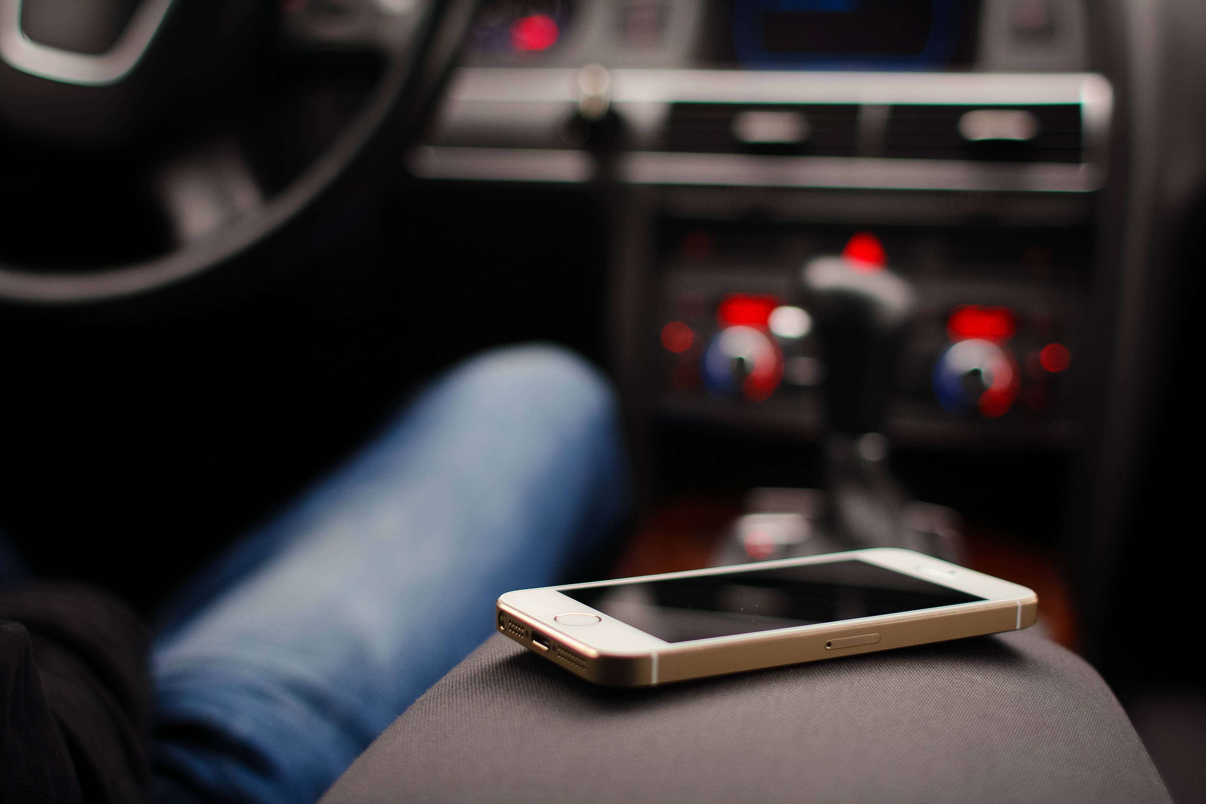 原版 iPhone 5 在車內 壁紙 — 一部金色智能手機放在車內一個人的大腿上，車內有紅色發光的刻度盤和模糊的儀表板。