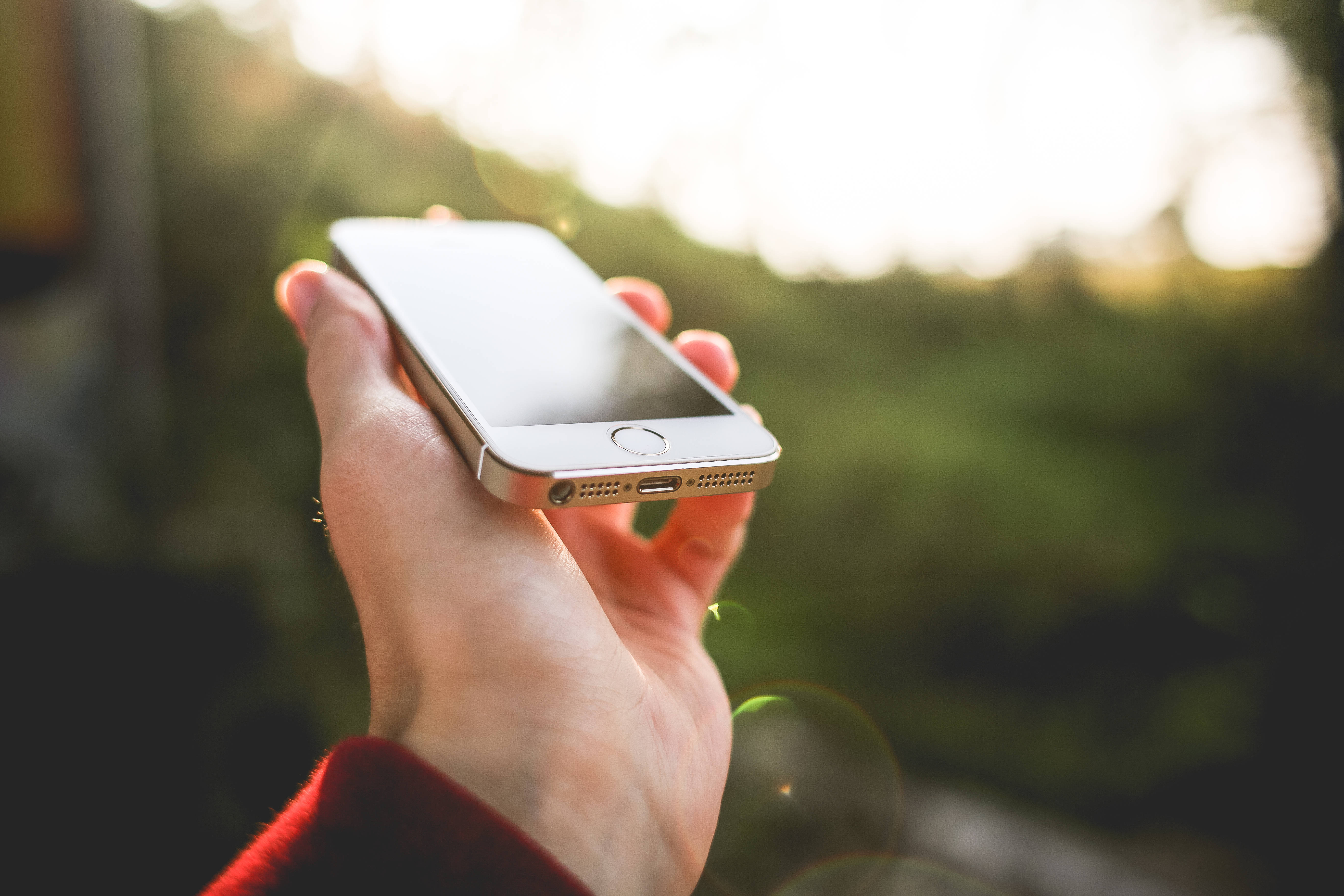 原裝 iPhone 5 聚焦 壁紙 — 一隻手拿著銀色智能手機，背景是陽光照射下模糊的綠色和鏡頭光暈。