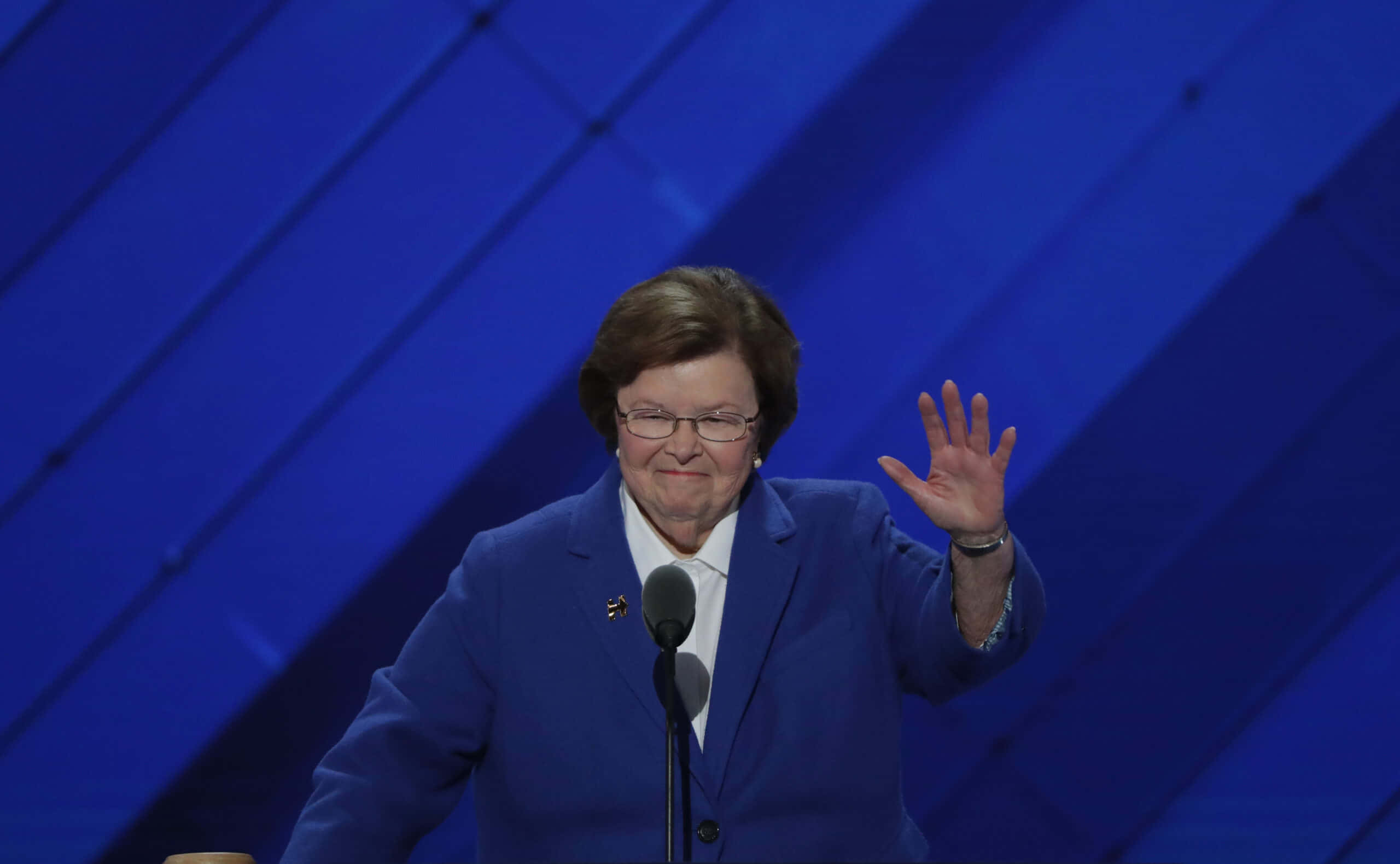 參議員 Barbara Mikulski 參與社區活動 壁紙 — 一位身穿藍色西裝的女性站在講台上，在幾何藍色背景前揮手。