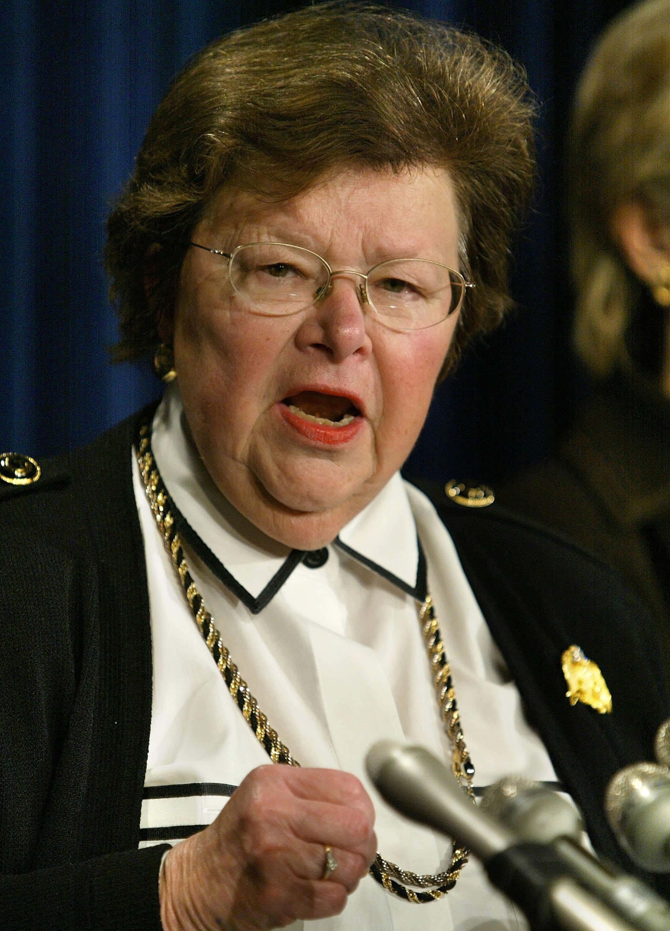 參議員 Barbara Mikulski 發表演講 壁紙 — 一位年長女性穿著正式制服，戴著金鏈和眼鏡，在藍色窗簾背景下對著麥克風熱情發言。