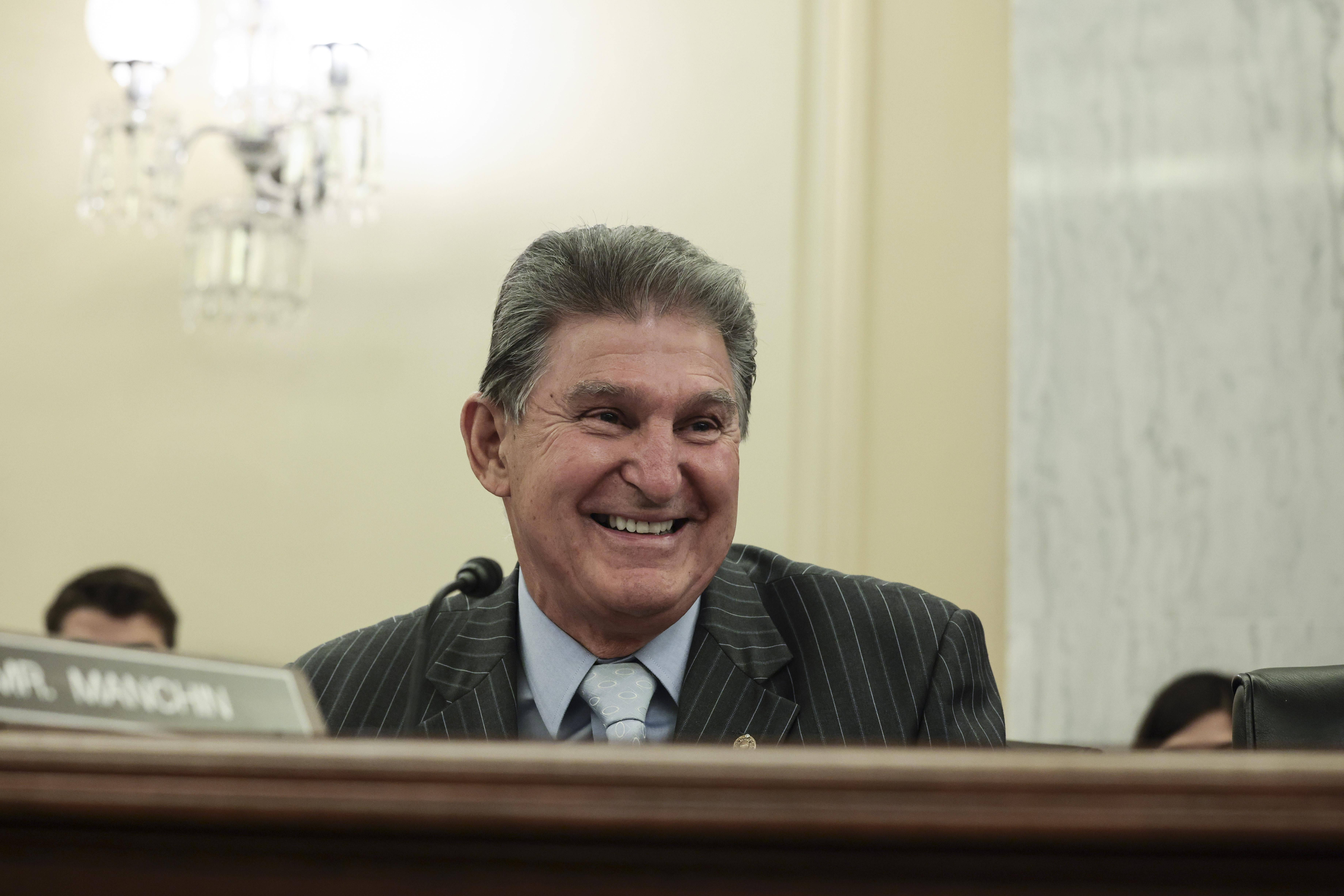 參議員 Joe Manchin 的大笑 壁紙 — 一位微笑嘅男士穿著條紋西裝，喺一個有吊燈嘅正式聆訊室入面，坐喺木枱前開懷大笑。