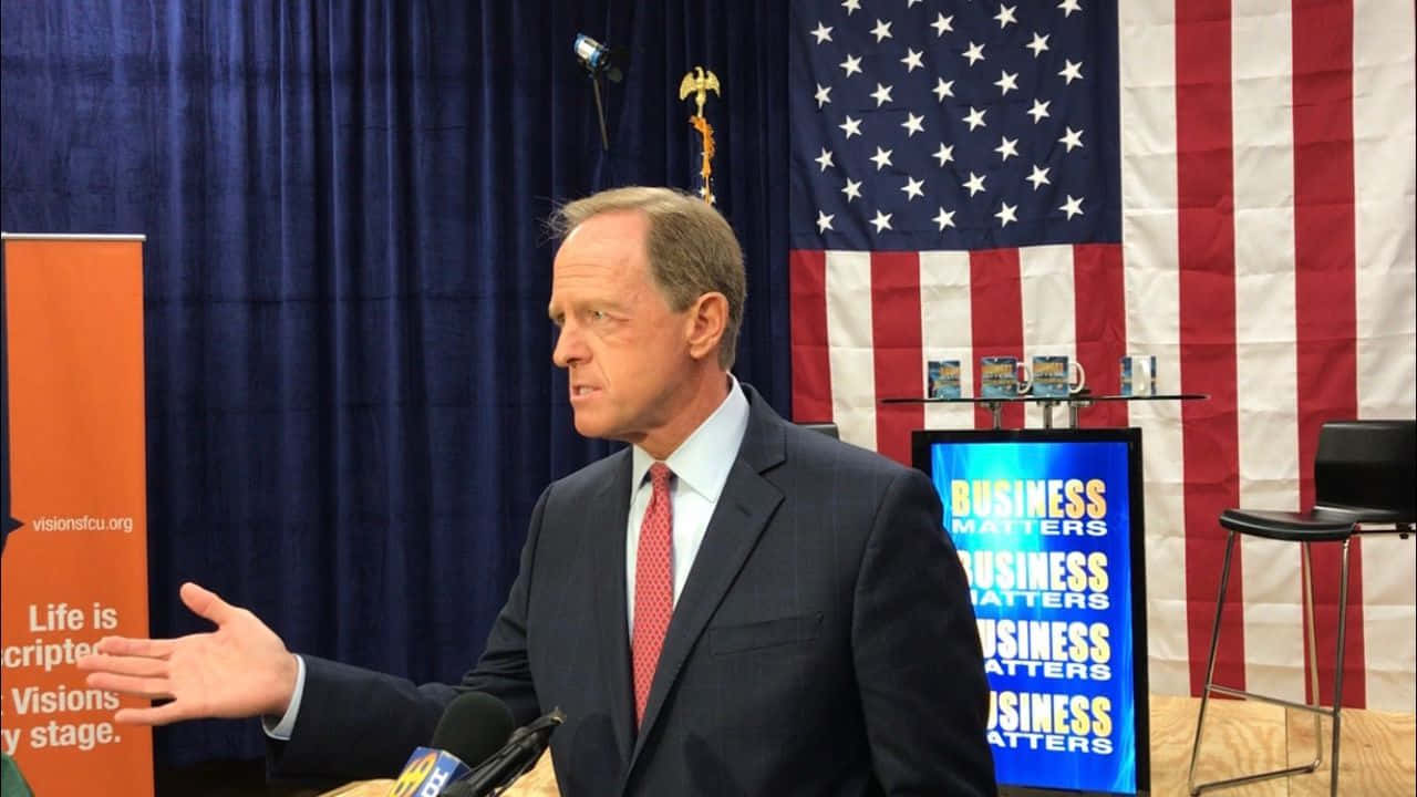 參議員Pat Toomey在商業活動中發言 壁紙 — 一名身穿西裝的男子在講台上說話時做手勢，身後有美國國旗和寫有「Business Matters」的屏幕。