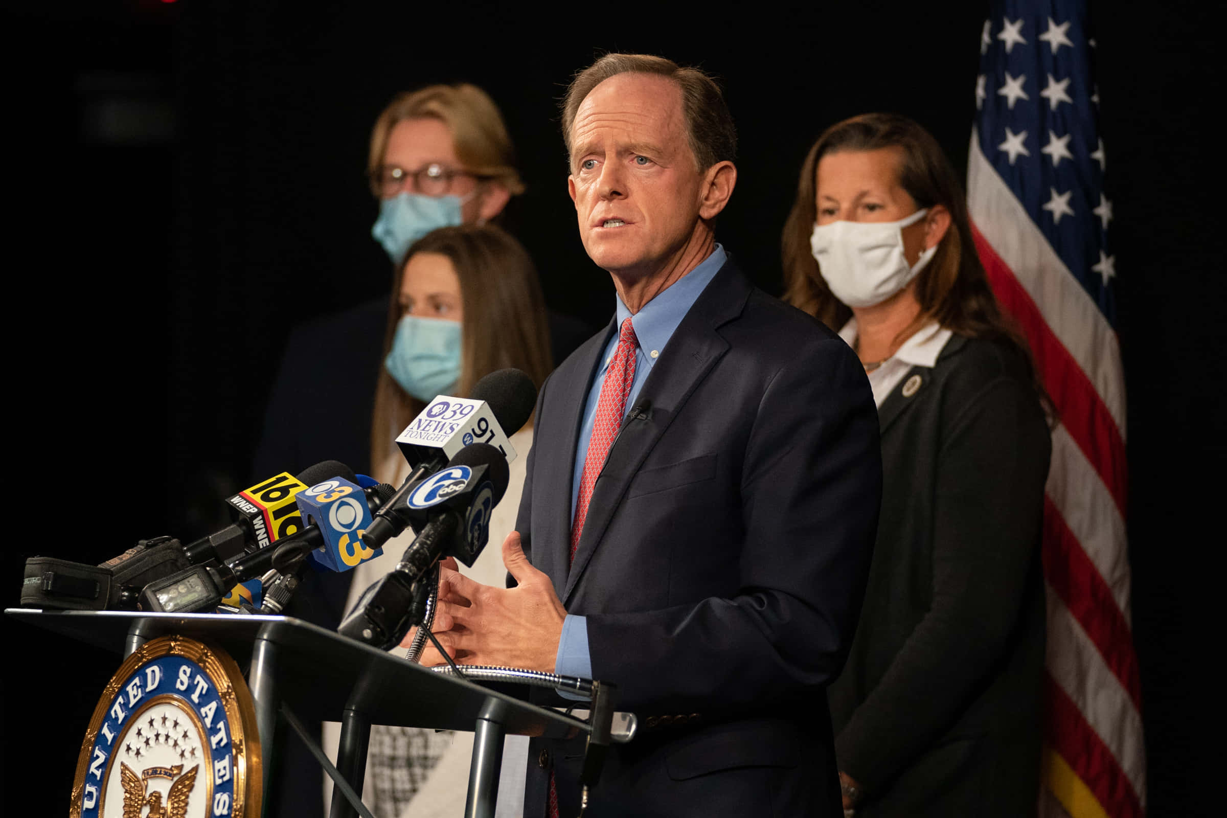 參議員Pat Toomey與媒體互動 壁紙 — 一名身穿西裝的男子在講台上發言，身後有麥克風和美國國旗，附近站著戴口罩的人。