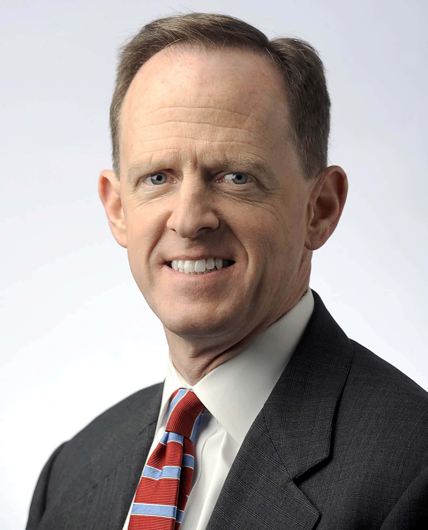 參議員帕特·圖米肖像照 壁紙 — 一位微笑的中年男子，身穿深色西裝，繫著紅藍條紋領帶，背景為純白色。