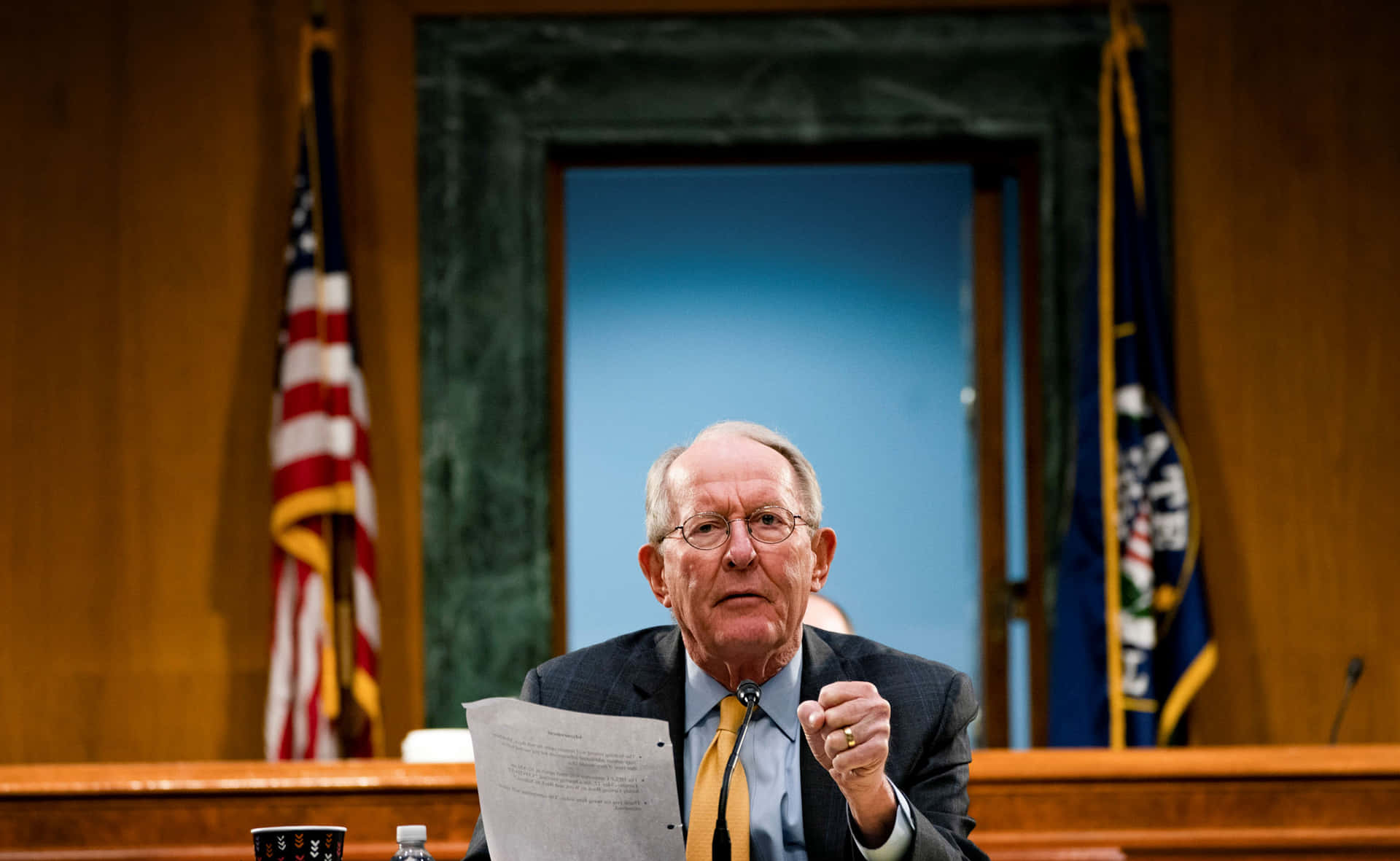 身穿西裝、戴黃色領帶的年長男性在政府聽證會上發言，身後有旗幟。