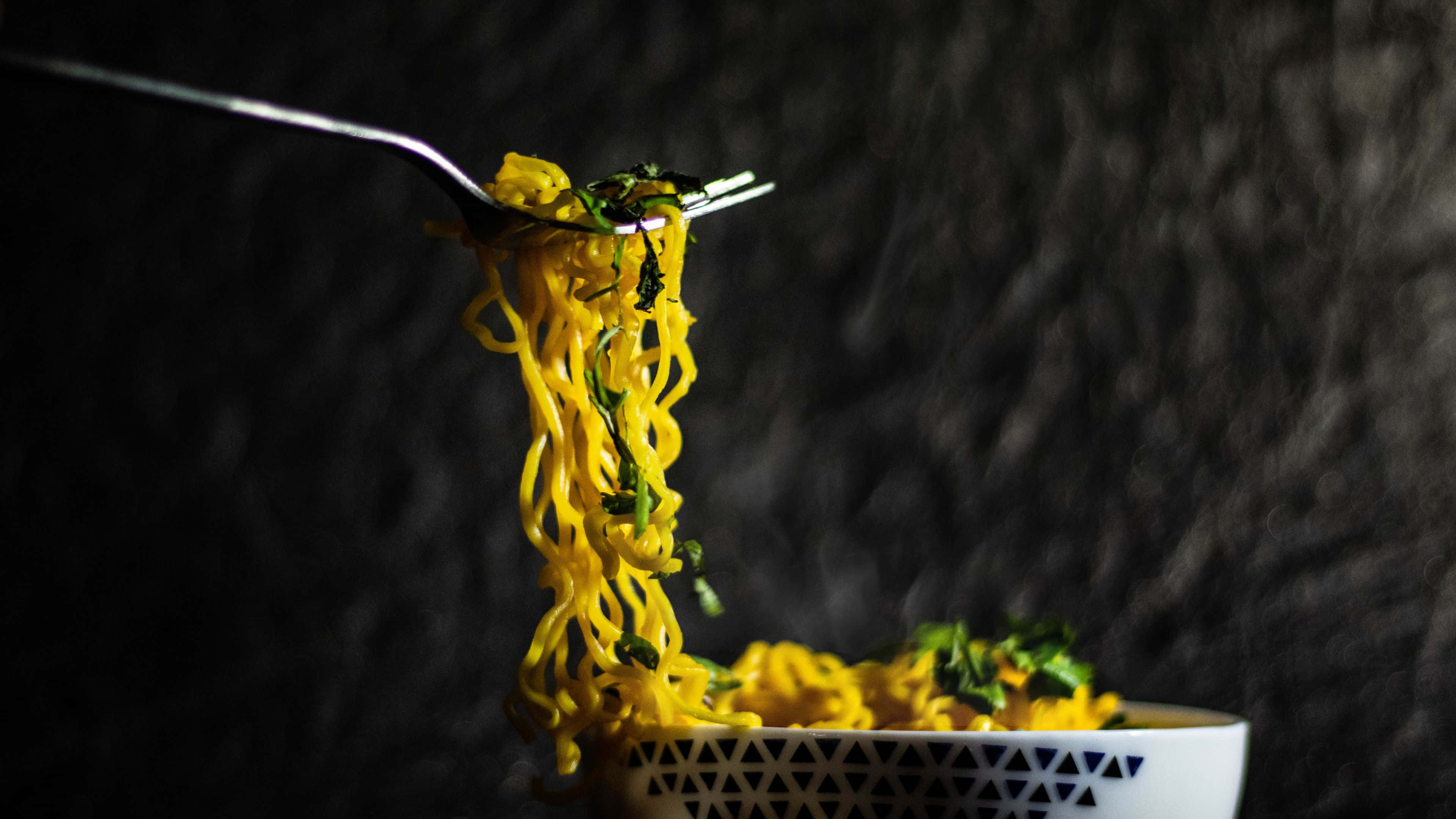 Noodles 桌布 壁紙 — 叉子從有圖案的碗中夾起冒著熱氣的黃色麵條和綠色香草，背景為深色紋理。
