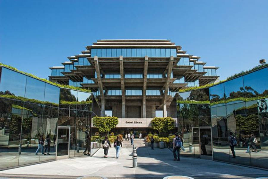 反射迷宮 UCSD 壁紙 — 現代玻璃和混凝土圖書館，階梯式屋頂反射藍天和綠意，人們走過入口。