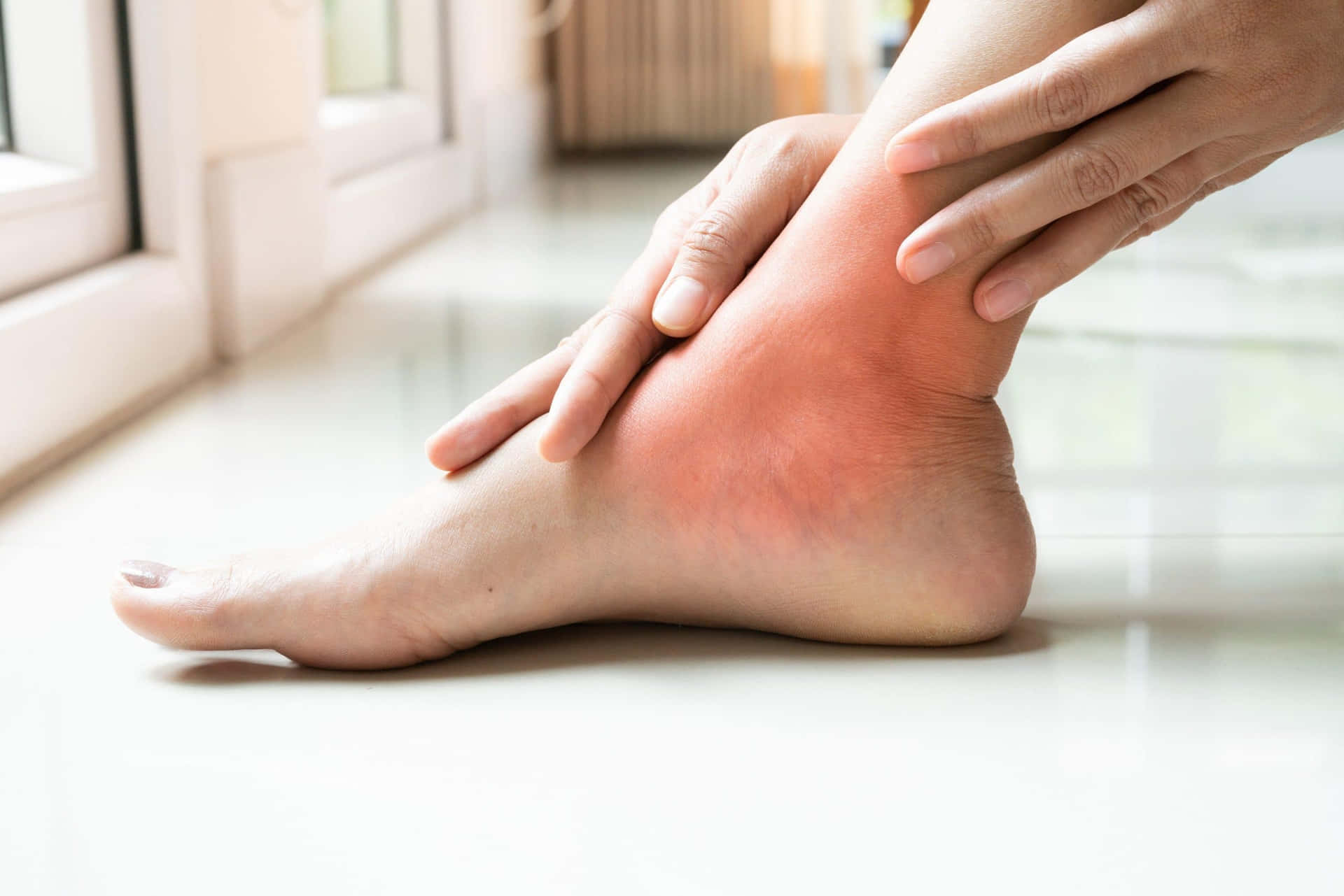 一個人在室內窗邊握住他們腫脹疼痛的腳踝。