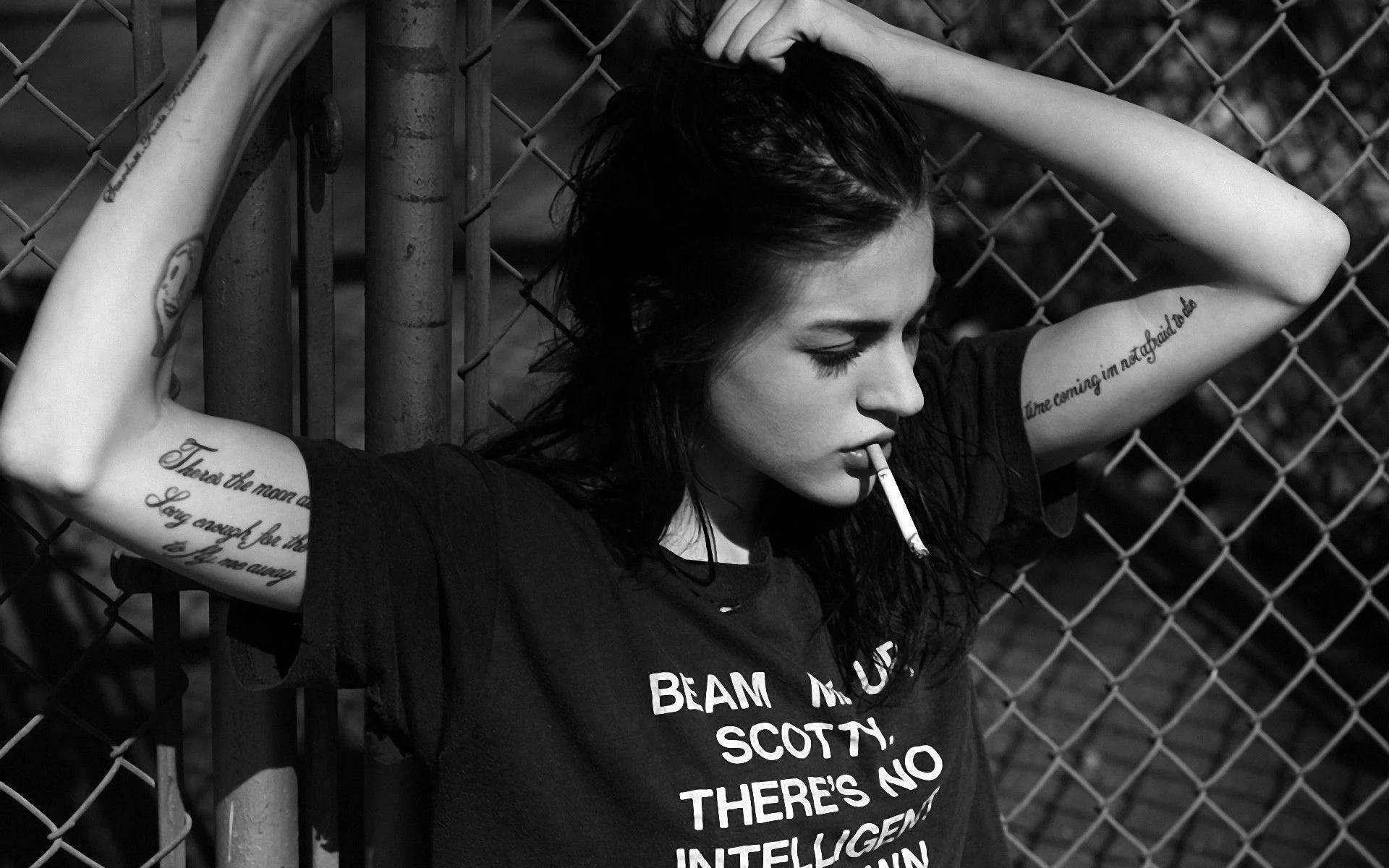叛逆精神的化身：抽菸的壞女孩 壁紙 — 一位有紋身的年輕女子靠在鐵絲網圍欄上抽煙，黑白照片。