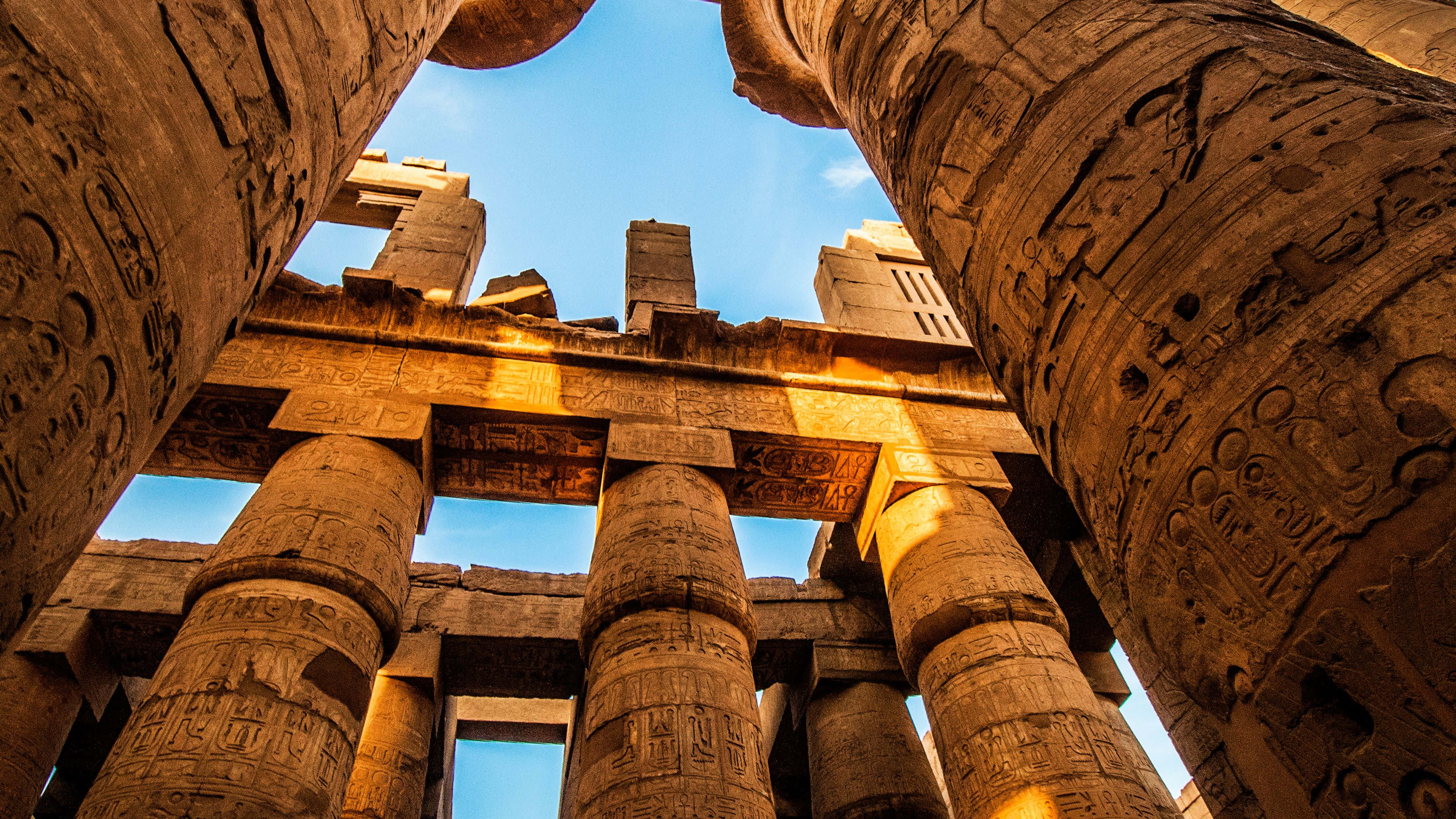 Ancient Egypt 桌布 壁紙 — 古埃及神廟的柱子雄偉地聳立在清澈的藍天下，雕刻著象形文字，沐浴在金色的陽光中。