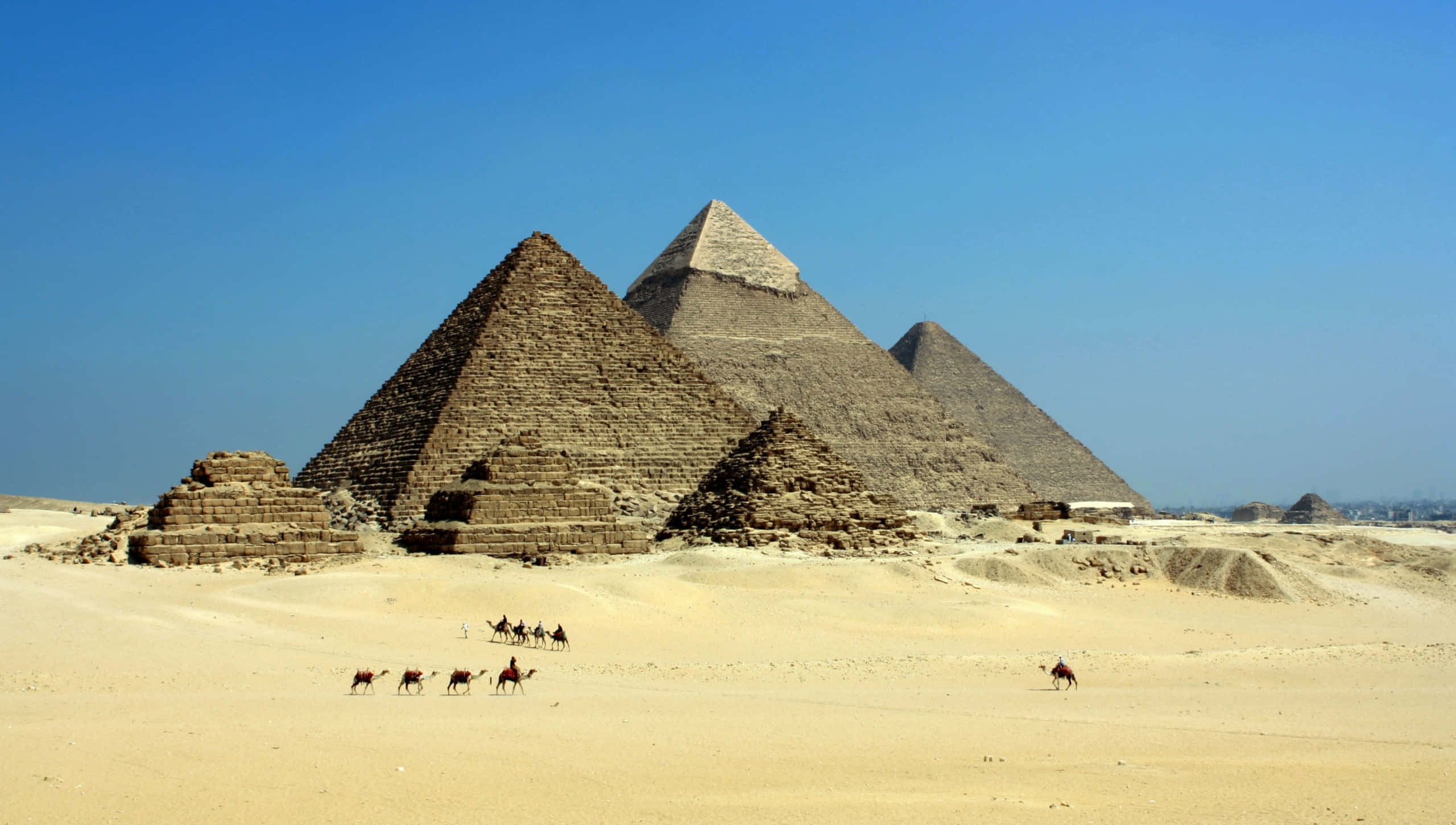 Ancient Egypt 桌布 壁紙 — 古埃及金字塔在金色沙漠中雄偉地聳立，清澈藍天下，駱駝和騎手穿越沙丘。