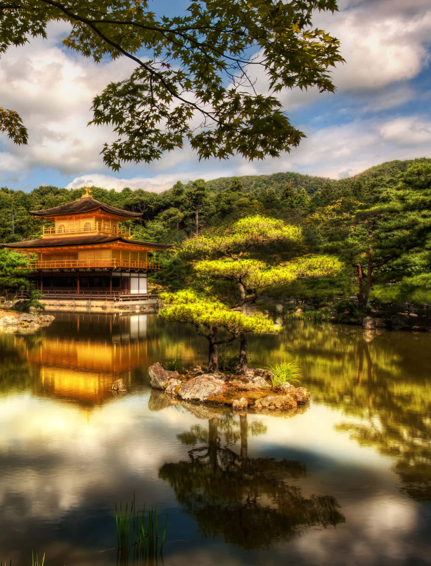 古寺靜立於傳統日式庭園之中 壁紙 — 金色嘅金閣寺喺部分多雲嘅天空下，喺平靜嘅水面中倒映，周圍係茂密嘅綠色樹木。
