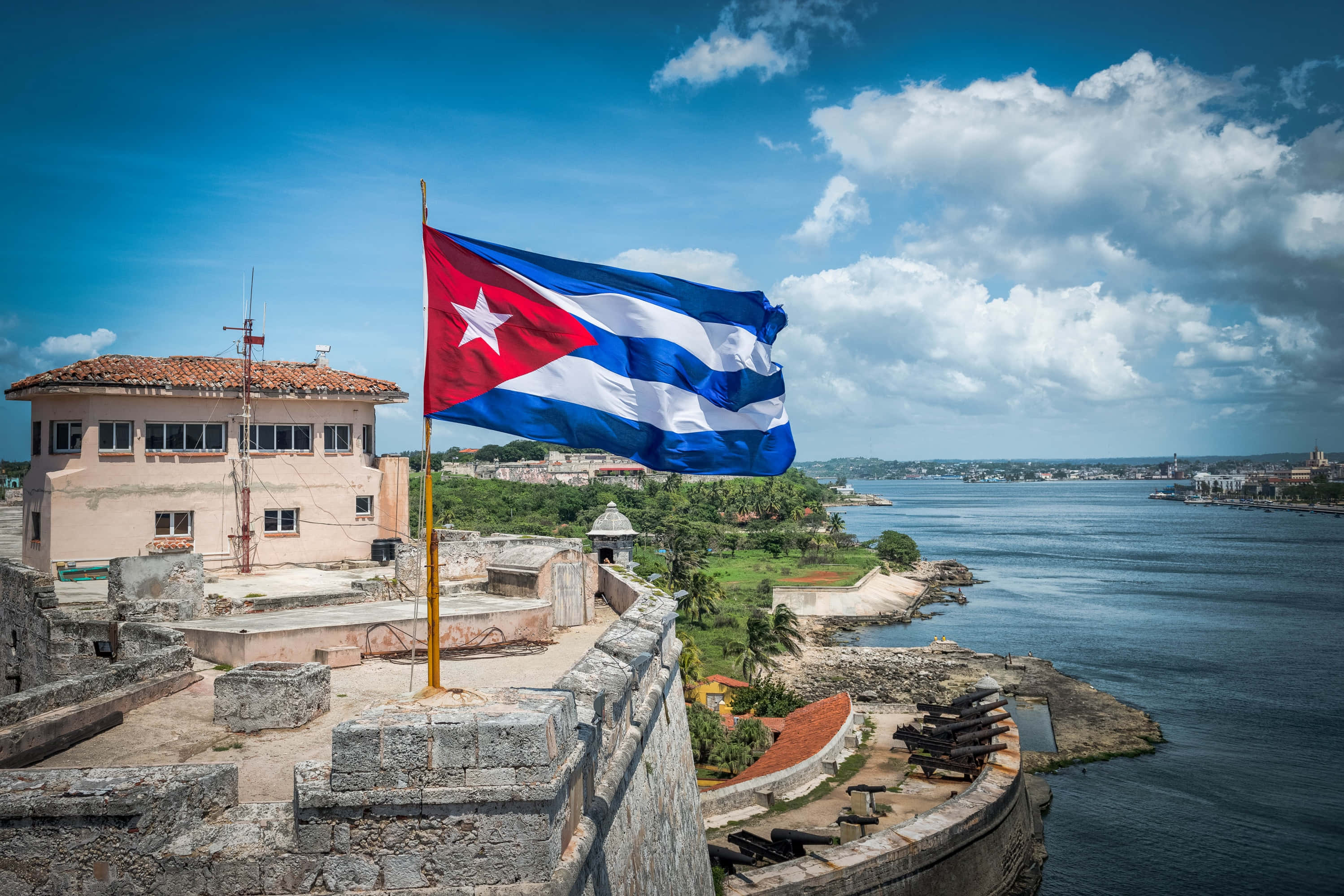 Cuba 桌布 壁紙 — 古巴國旗在歷史悠久的石堡壘頂部自豪地飄揚，俯瞰著沿海城市和碧綠的海水，天空湛藍明亮。