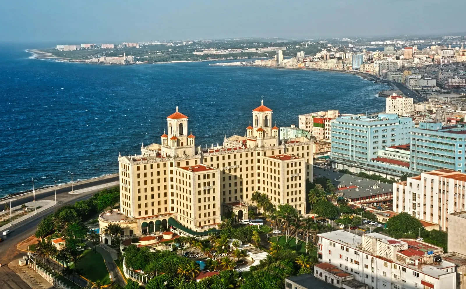 古巴翠綠鄉野上的美麗日落 壁紙 — 從高空俯瞰，一座米色大酒店，紅色屋頂，隱藏在棕櫚樹之間，旁邊是湛藍的海洋和廣闊的海濱城市。