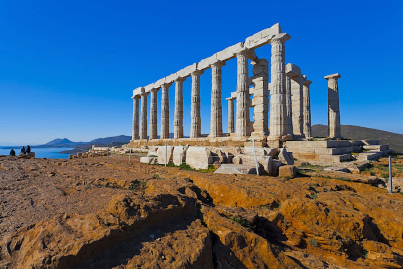 greek 桌布 壁紙 — 古希臘神廟遺址，高聳的柱子矗立在崎嶇的橙色懸崖上，俯瞰著晴朗天空下平靜的藍色大海。