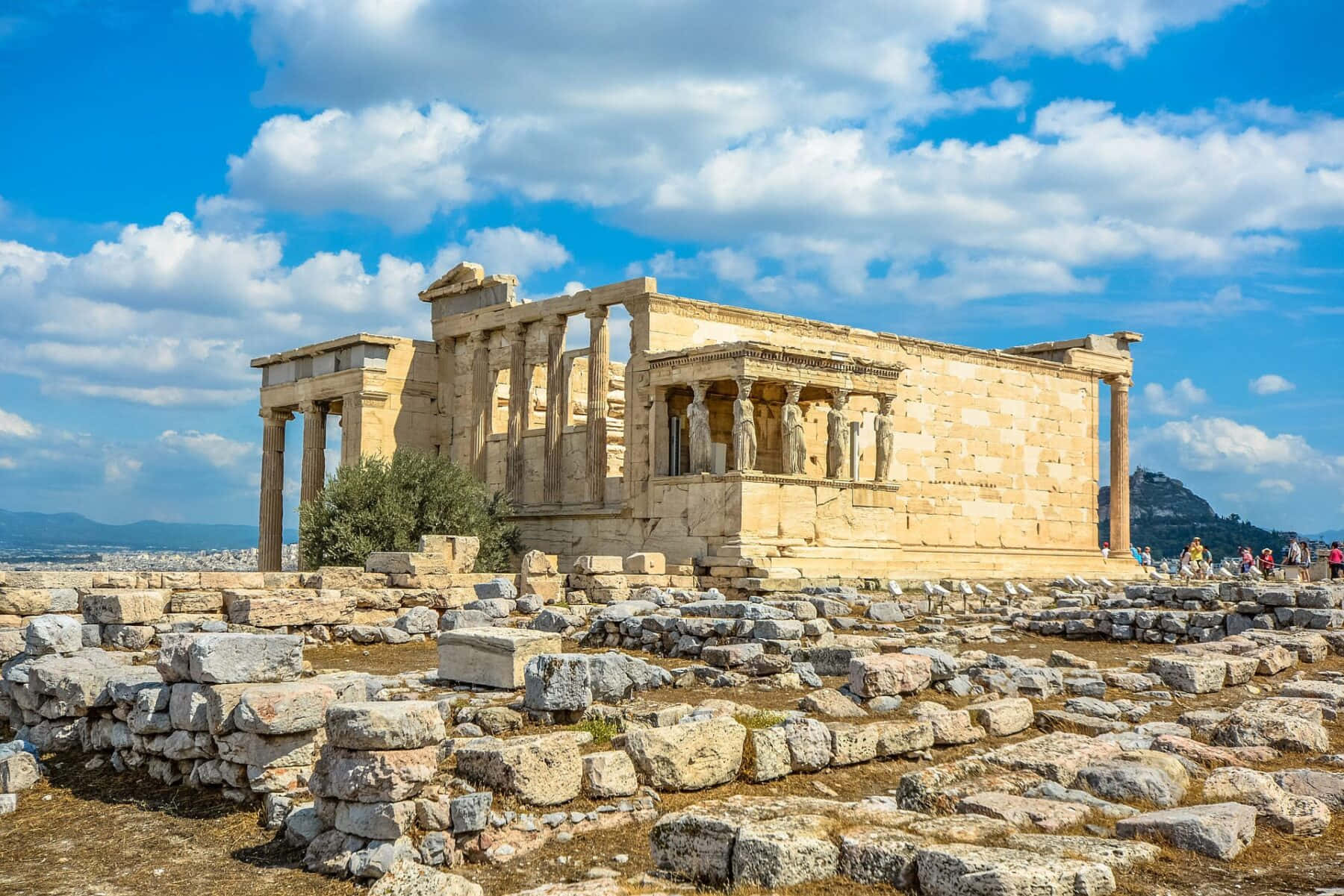 greek 桌布 壁紙 — 古老的石廟，帶有柱子和雕像，在明亮的藍天和蓬鬆的白雲下雄偉地矗立。
