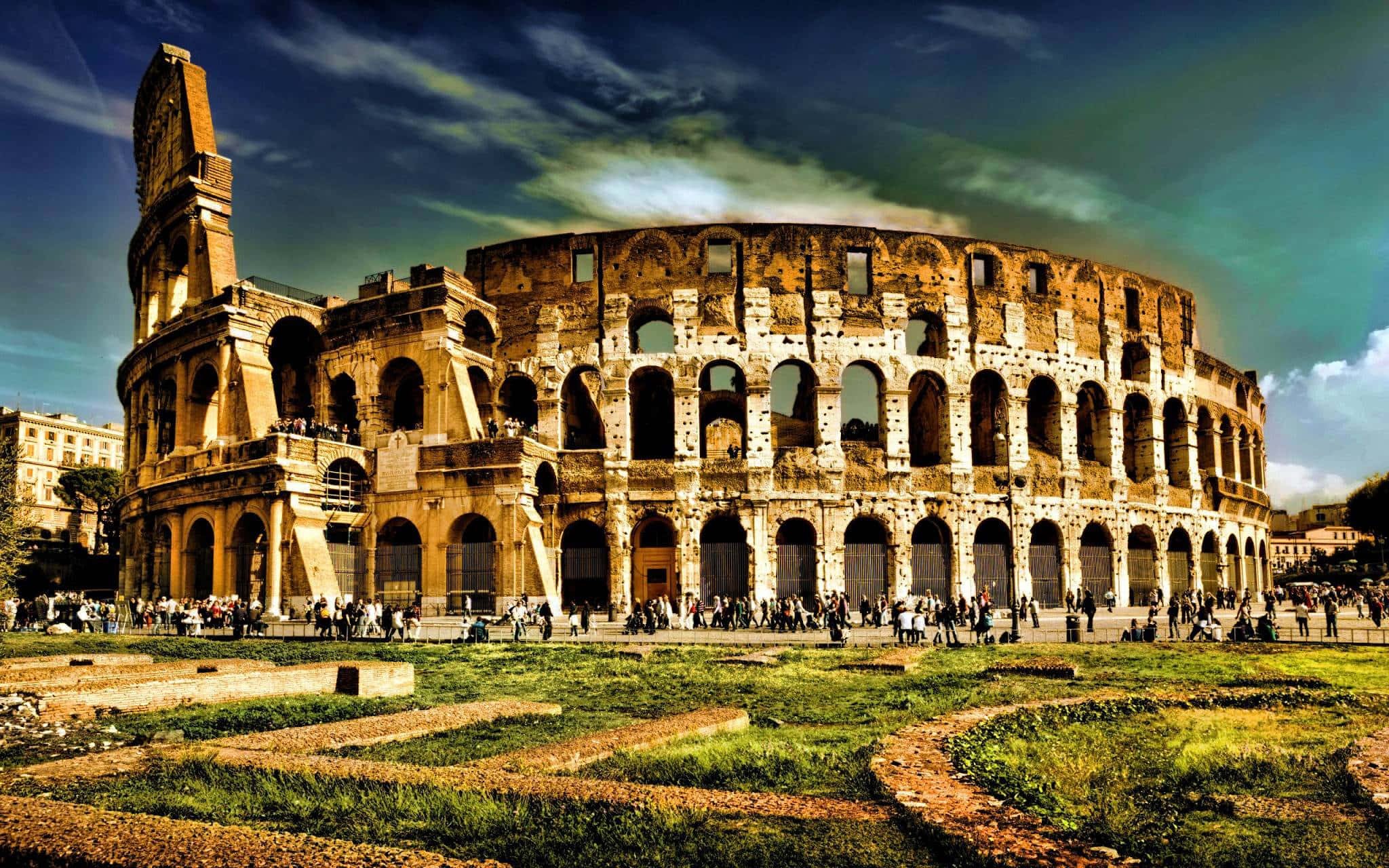 Rome 桌布 壁紙 — 古老的羅馬鬥獸場在戲劇性的天空下雄偉地矗立，遊客正在探索其歷史遺跡和周圍的草地。
