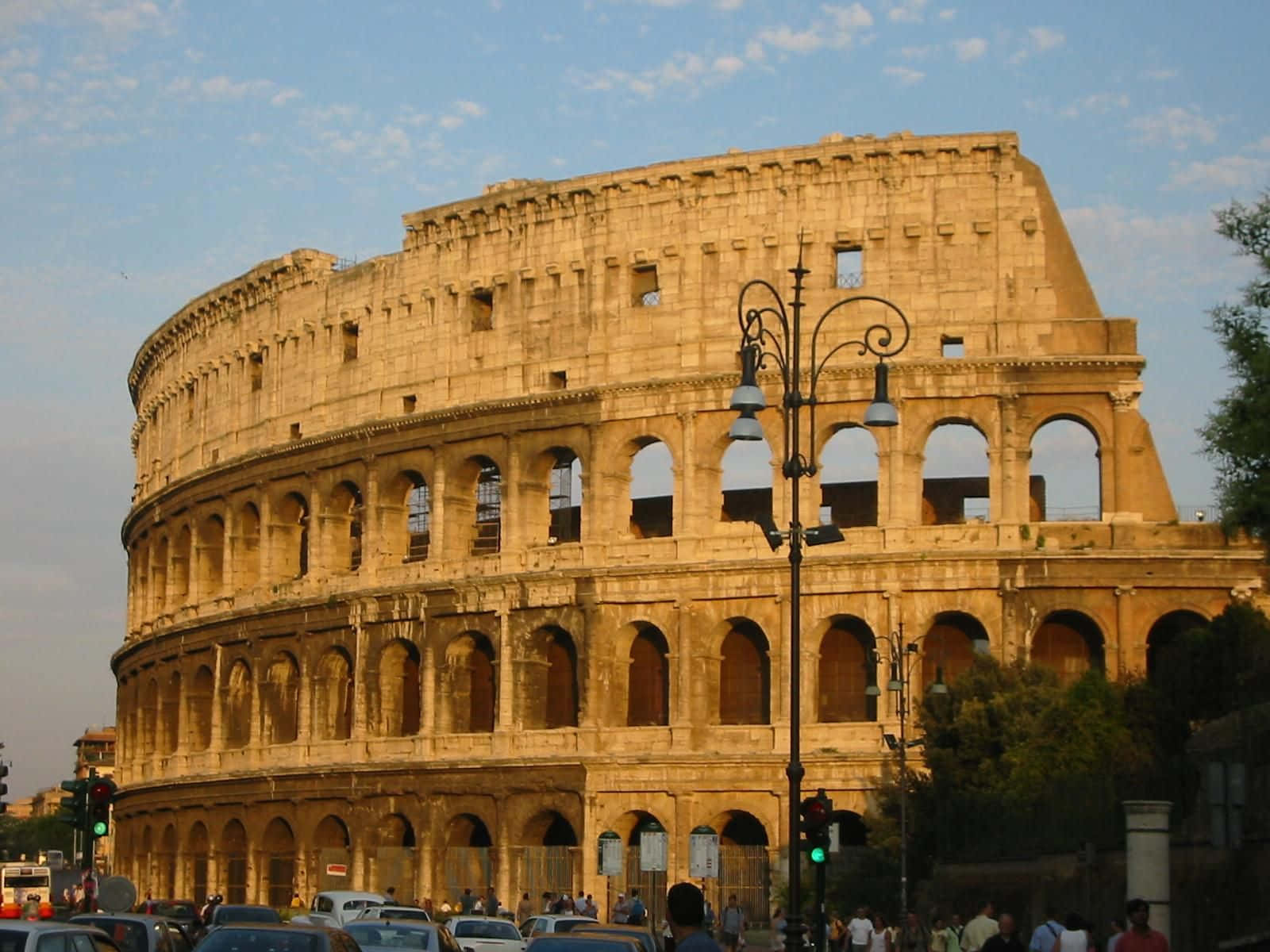 Roman 桌布 壁紙 — 古老的羅馬鬥獸場在柔和藍天與零星白雲下巍然聳立，前景街道上汽車和行人往來穿梭。