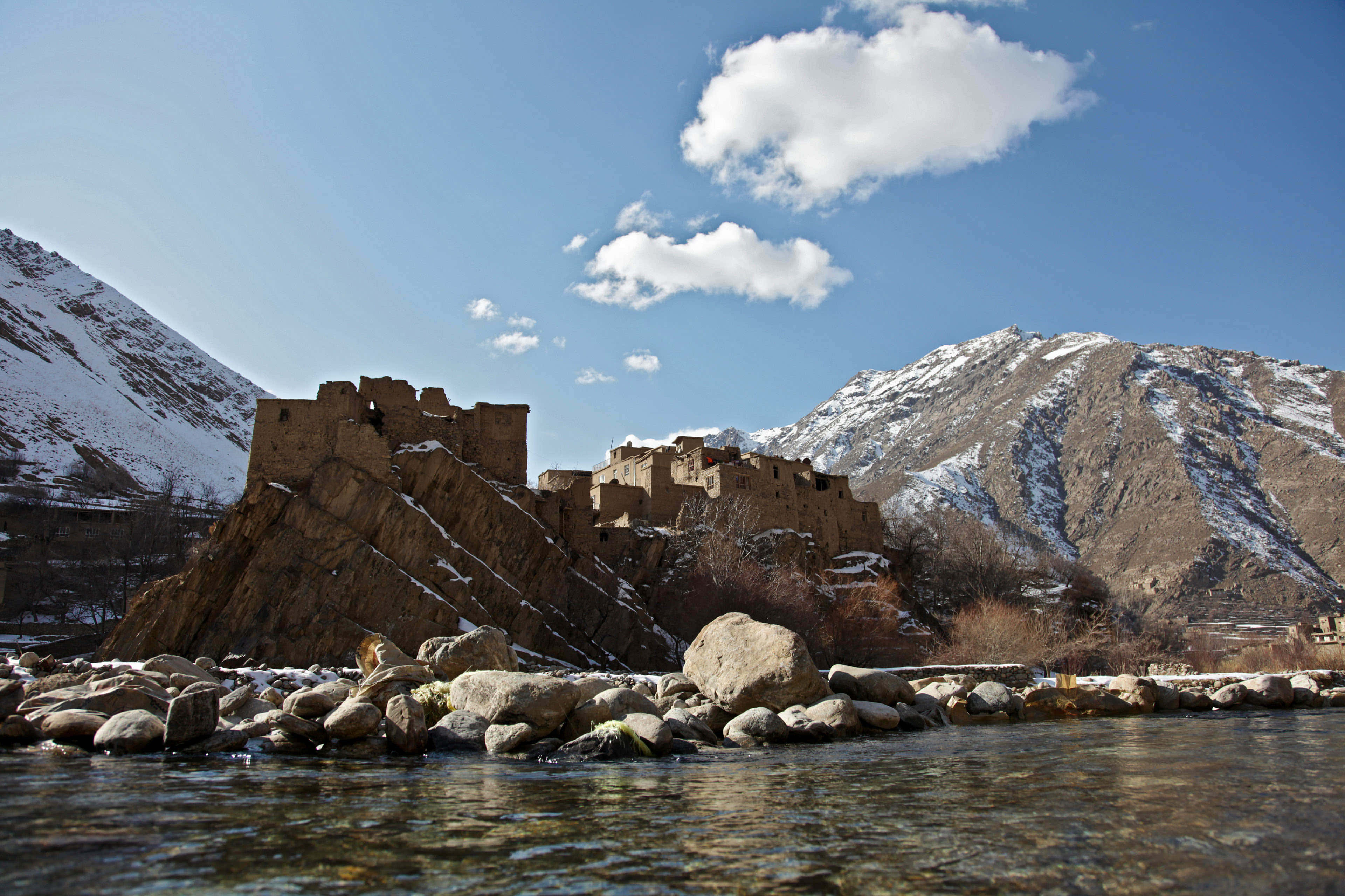 Afghanistan 桌布 壁紙 — 古老石堡坐落於岩石河岸上，白雪覆蓋的山脈在明亮的藍天下聳立。