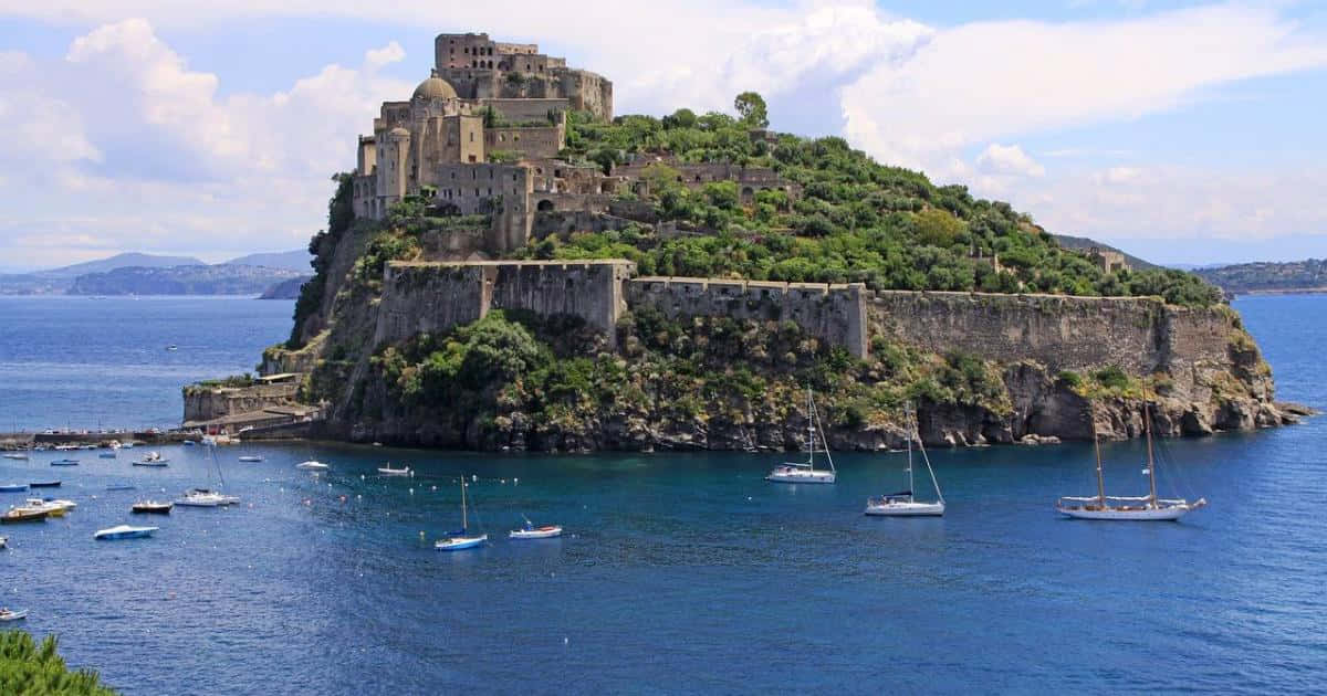 Italian Island 桌布 壁紙 — 古老石堡坐落於鬱鬱蔥蔥的懸崖上，俯瞰著點綴著帆船的蔚藍大海，天空部分多雲。