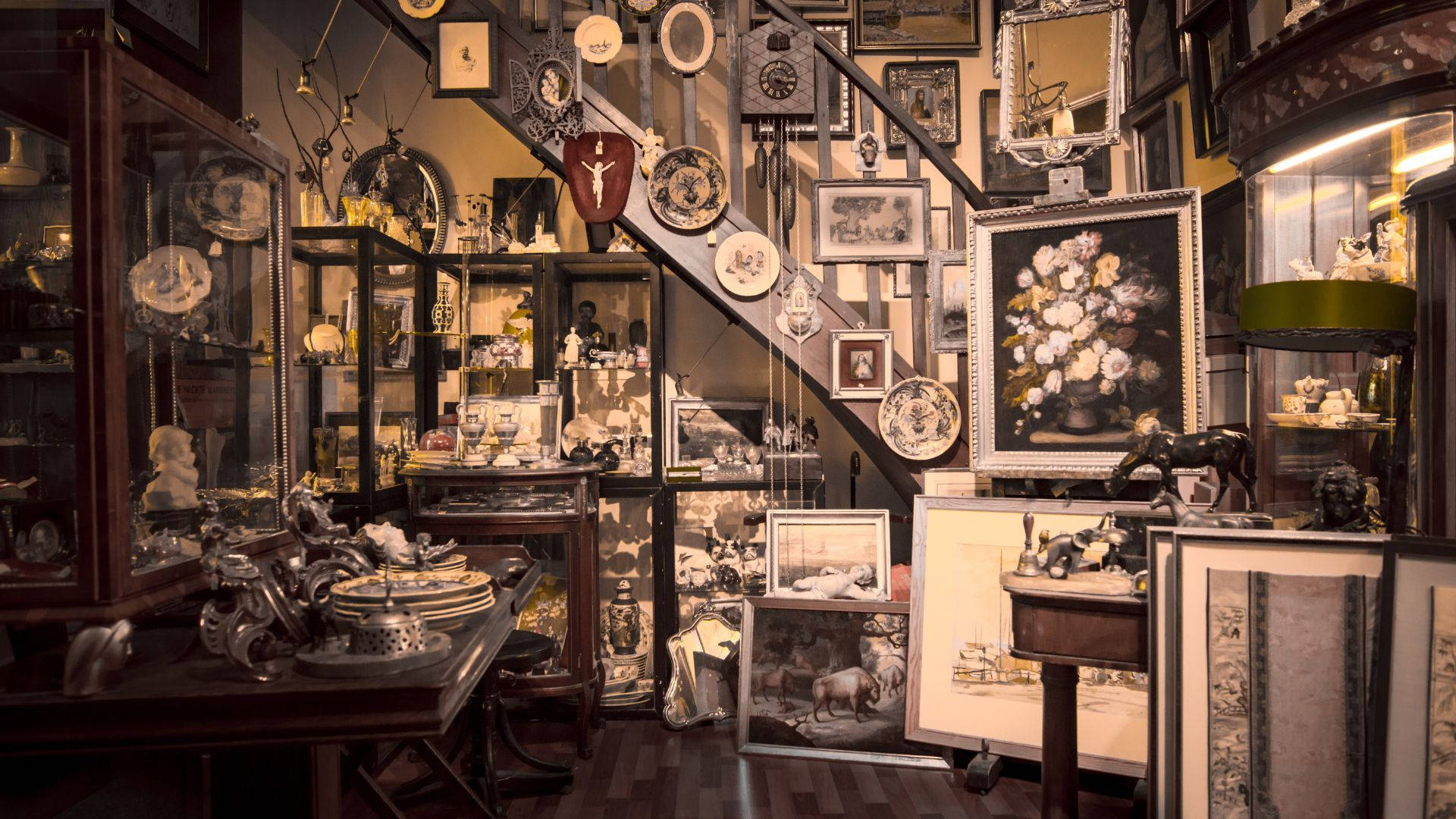 古董店與尼克納克餐廳 壁紙 — 一間燈光溫暖的古董店，中央樓梯周圍擺滿玻璃陳列櫃、復古畫作和華麗裝飾。