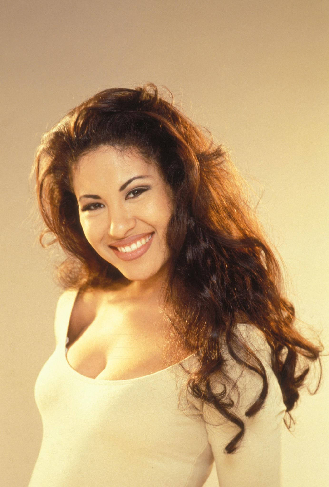 可愛 Selena Quintanilla 壁紙 — 一位容光煥發、擁有豐盈棕色捲髮的女性，在柔和的米色工作室背景前溫暖地微笑。