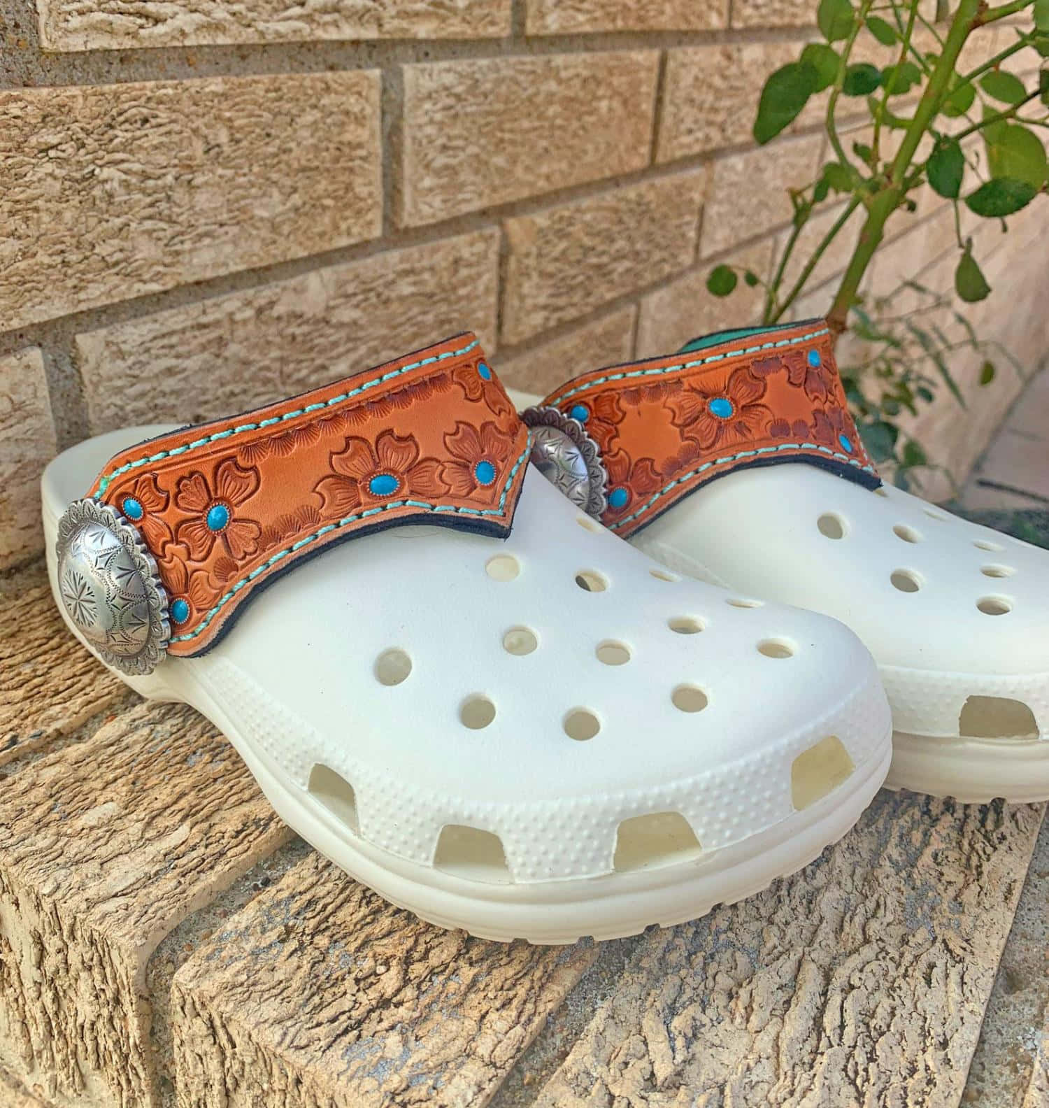 白色Crocs鞋，帶有裝飾性皮帶和綠松石，放在石面上。
