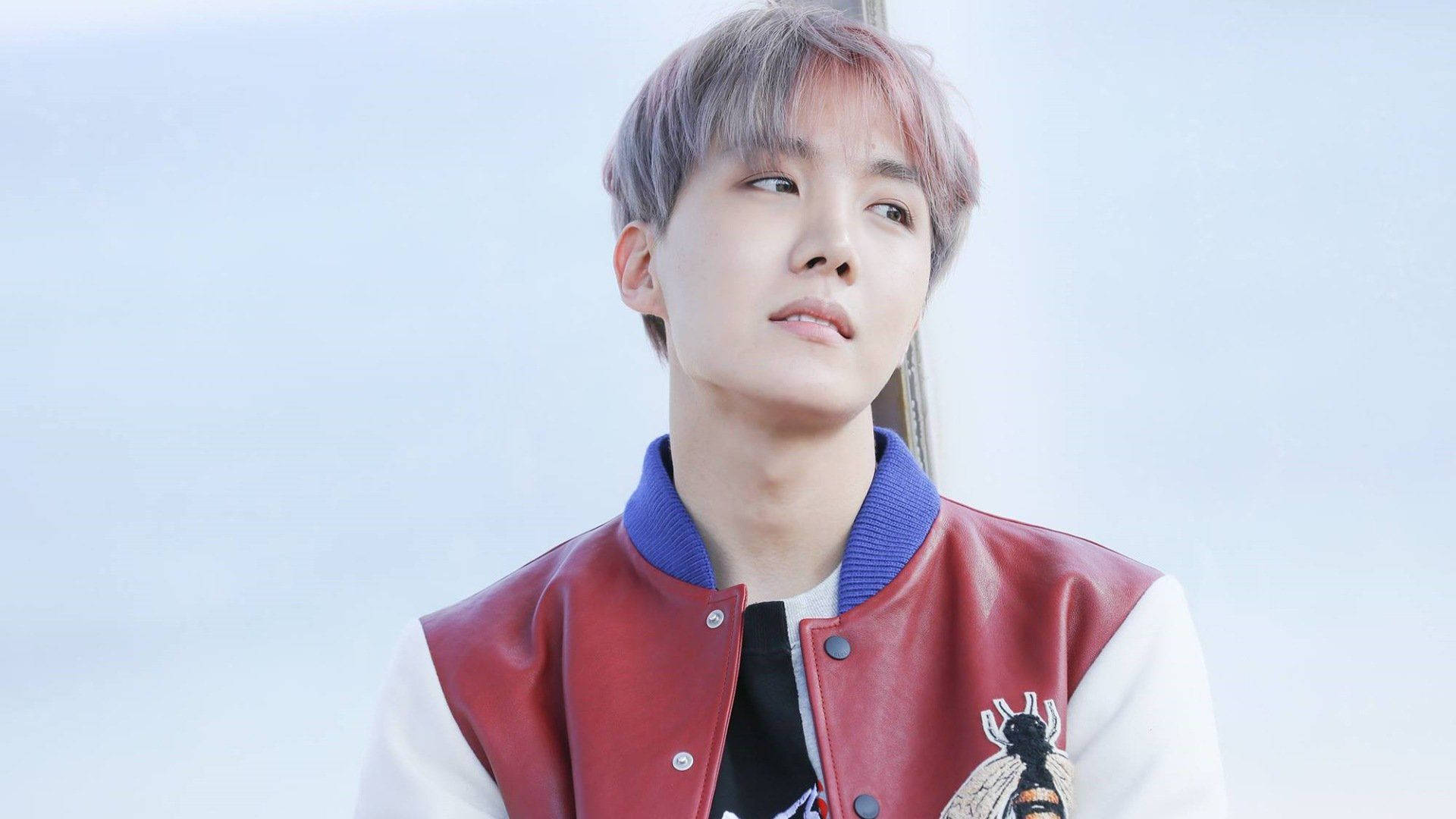 可愛BTS J-Hope穿紅白夾克 壁紙 — 一位擁有粉紅色頭髮的年輕男子，穿著紅白相間的校隊夾克，背景是柔和的藍天。