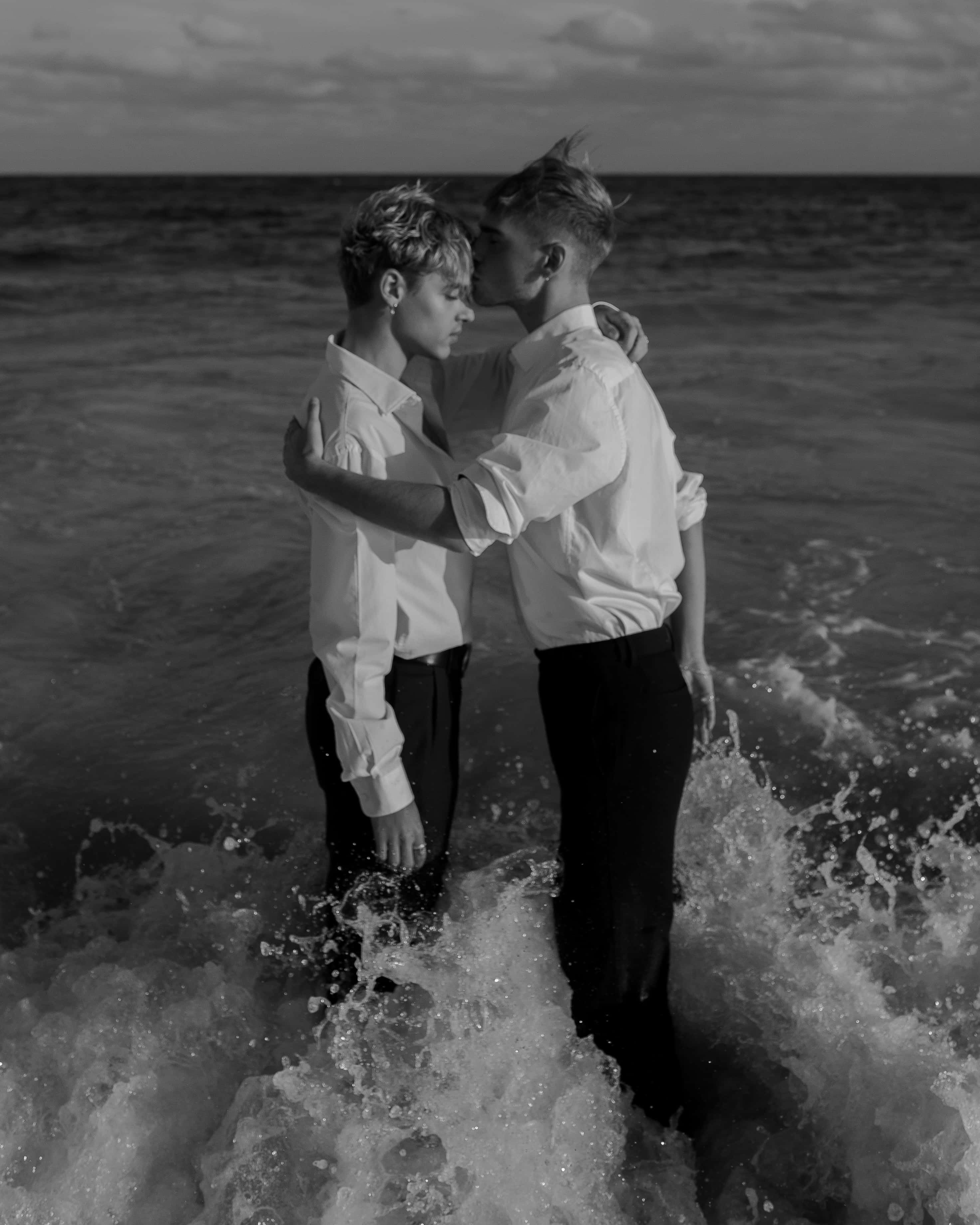 可愛LGBT情侶復古單色 壁紙 — 兩個身穿白恤衫的男人在黃昏的海浪中溫柔擁抱，黑白照片。