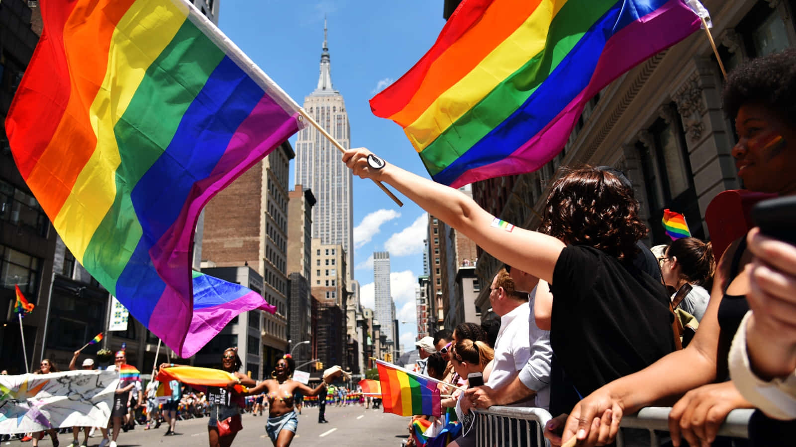 可愛LGBT旗幟紐約市驕傲遊行 壁紙 — 在驕傲遊行期間，鮮豔的彩虹旗高高飄揚在繁忙的城市街道上方，背景是帝國大廈。
