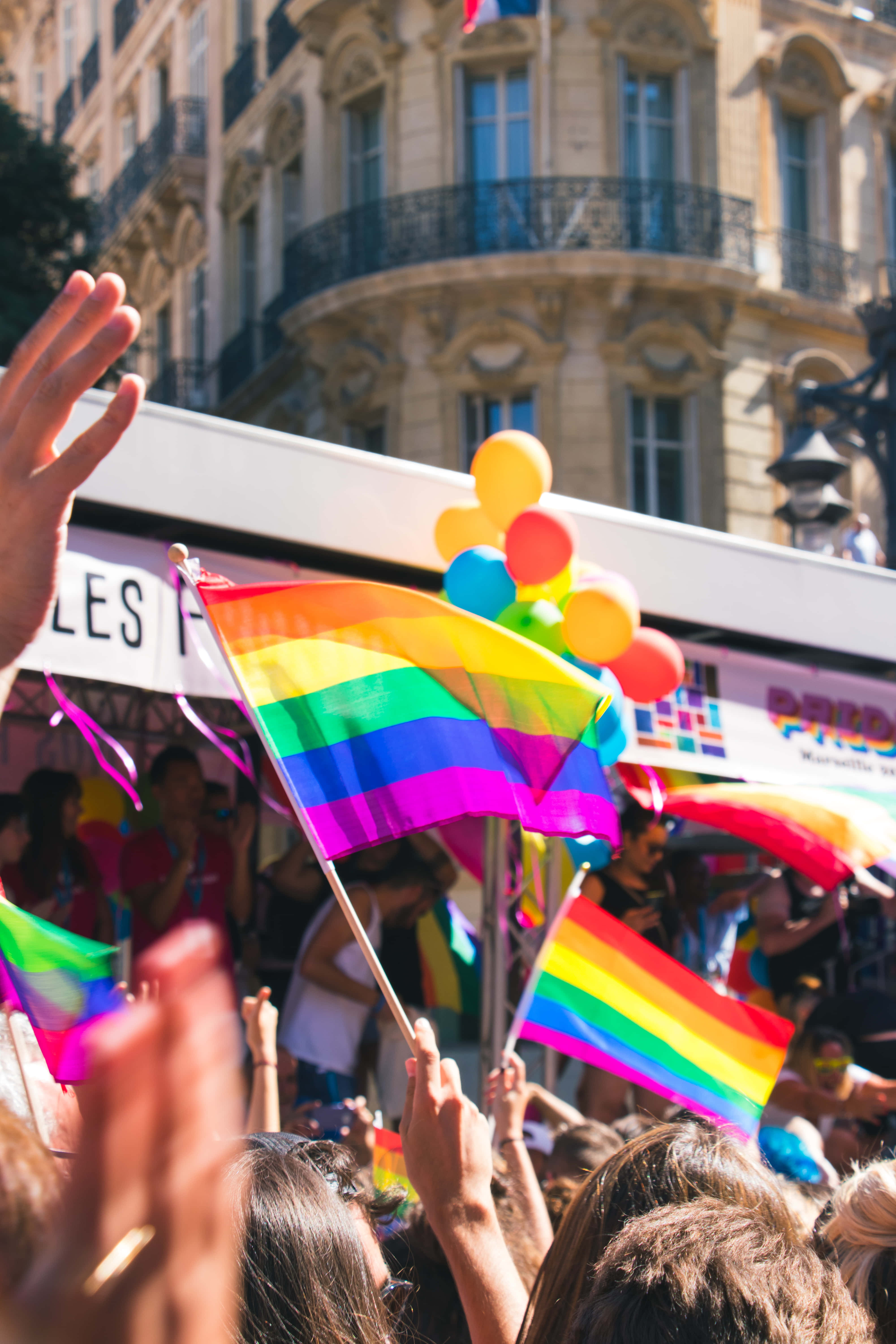 可愛LGBT旗幟驕傲遊行 壁紙 — 在歐洲城市街道上，陽光明媚的驕傲遊行中，充滿活力的人群揮舞著彩虹旗和彩色氣球。