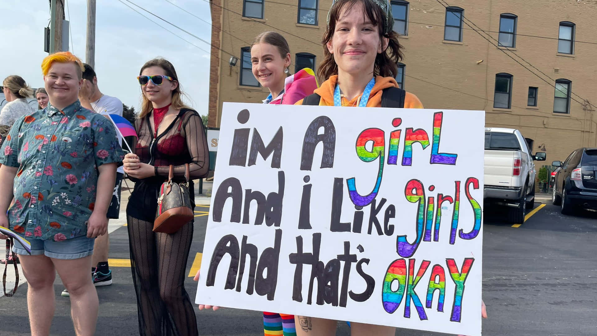 可愛LGBT活動家百老匯大道攝影 壁紙 — 一位年輕人在戶外驕傲活動中微笑著舉起彩虹色的標語牌，上面寫著「我是女孩，我喜歡女孩，這沒關係」。