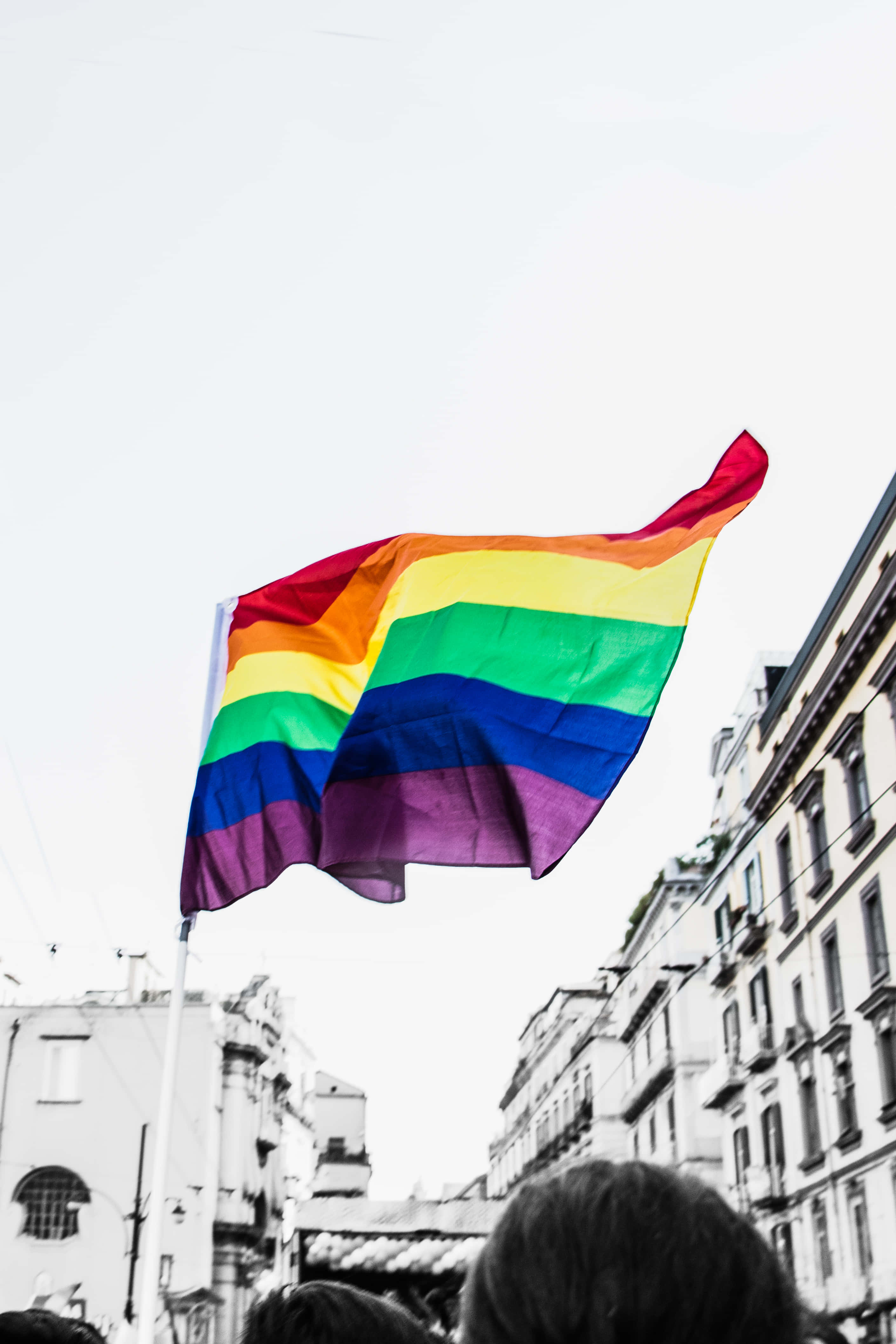 可愛LGBT驕傲旗2018年攝影 壁紙 — 一面鮮豔的彩虹旗在歷史建築林立的繁華城市街道上空，對著明亮的天空自豪地飄揚。
