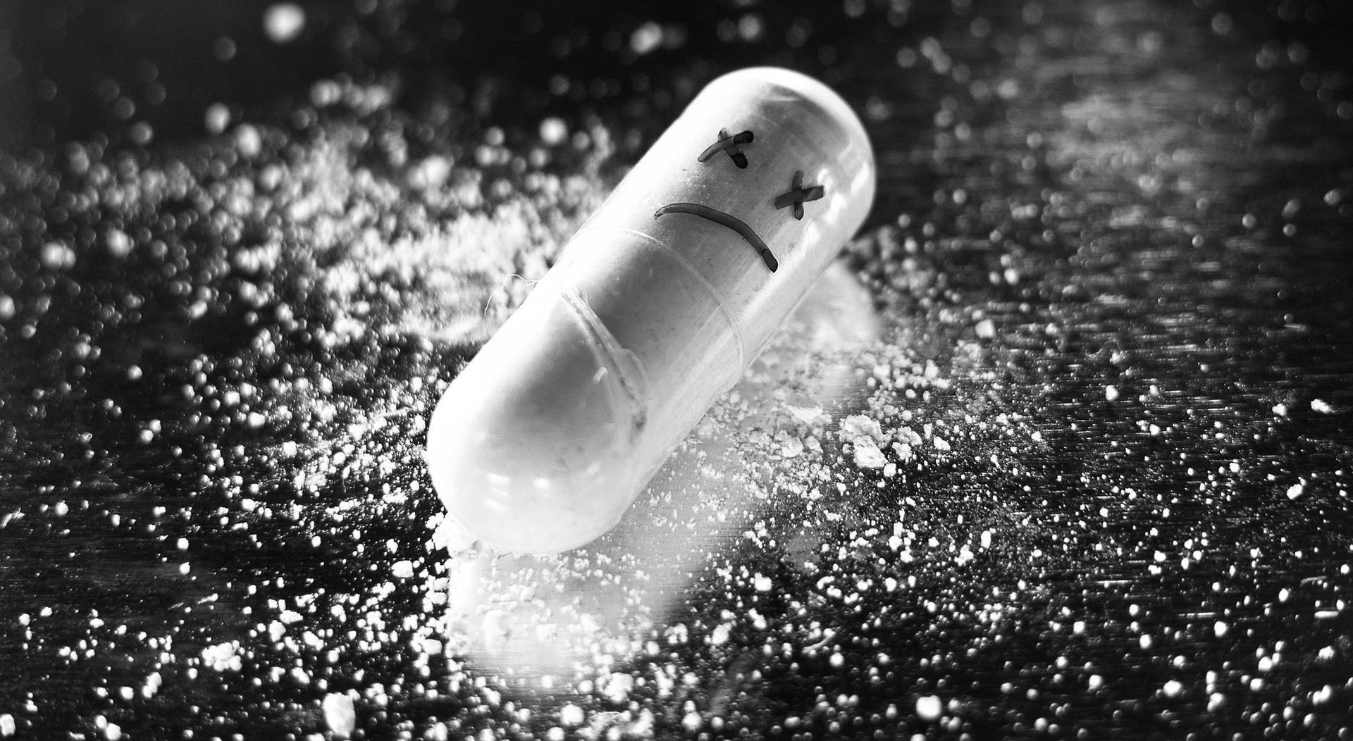 可愛傷心藥丸 壁紙 — 一張黑白照片，顯示一顆悲傷表情的藥丸躺在黑暗表面上，周圍散落著粉末。
