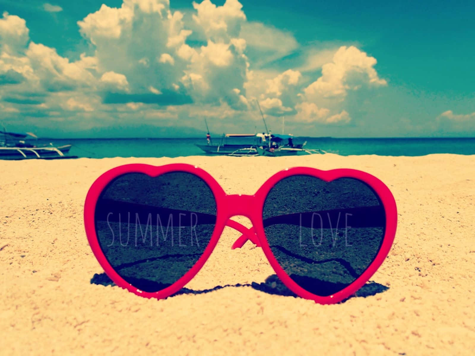 可愛夏日桌面心形眼鏡 壁紙 — 心形粉紅色太陽眼鏡，上面有'SUMMER'和'LOVE'字樣，放在沙灘上，背景有船隻和多雲的天空。