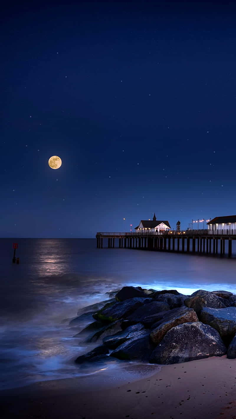可愛夜間海灘手機壁紙 壁紙 — 一個寧靜的月夜海灘，滿月高掛在星空之上，一條亮著燈的碼頭延伸至平靜的水面。