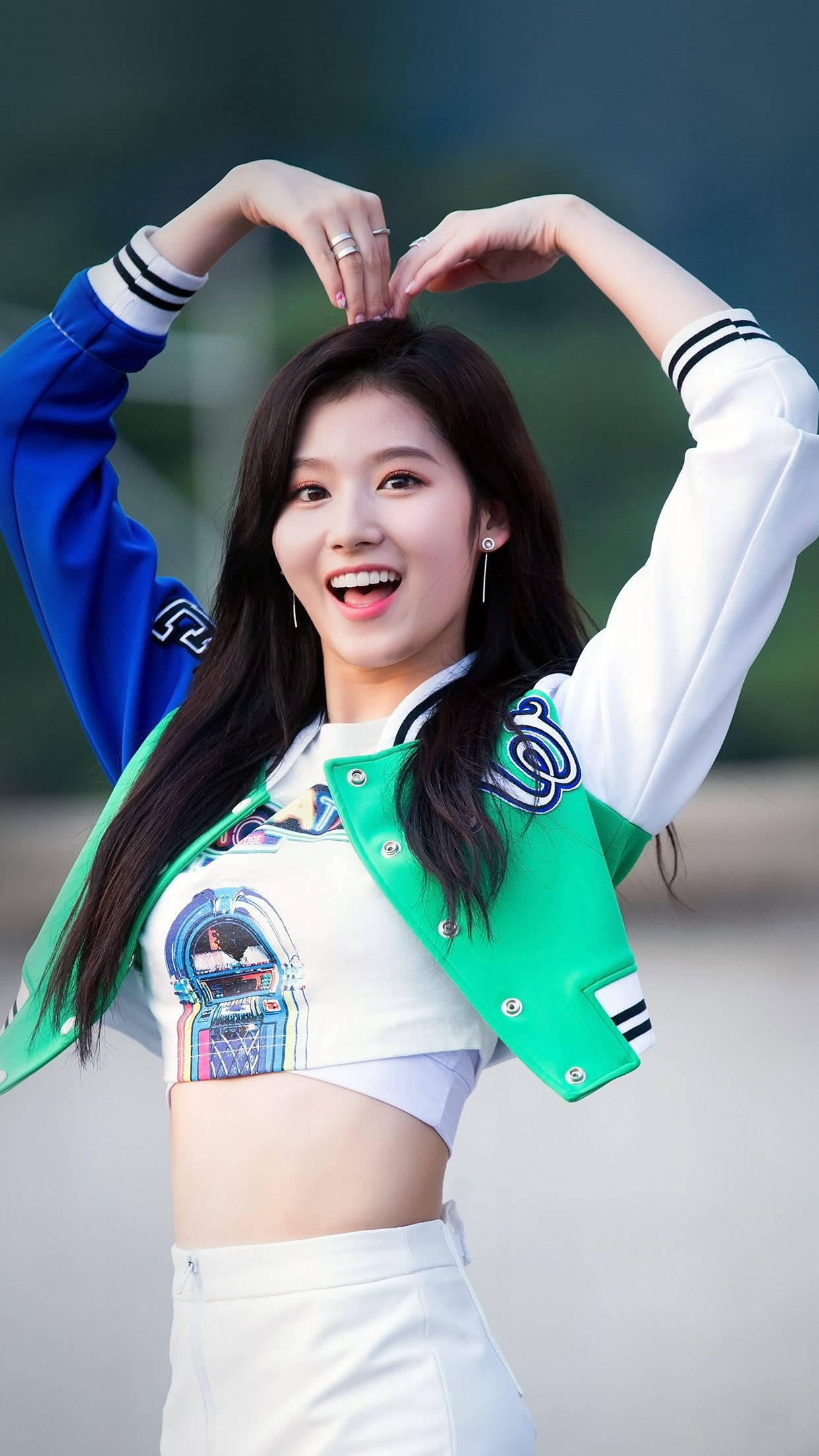 可愛女孩 K-pop Sana 壁紙 — 一位身穿色彩繽紛大學外套的快樂年輕女性，在戶外燦爛微笑，雙手在頭頂比出心形。