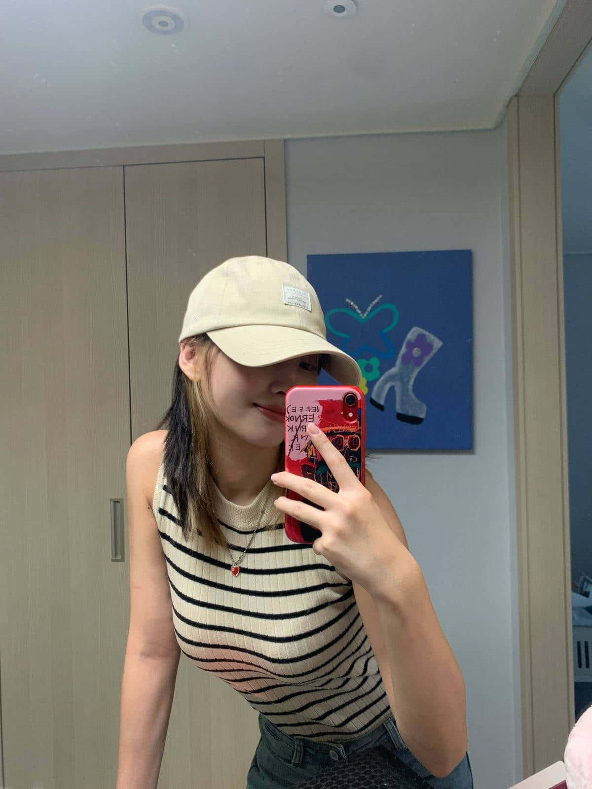 可愛女孩戴帽鏡頭自拍圖 壁紙 — 一位戴米色帽子和條紋上衣的年輕女性在現代房間裡用紅色手機對著鏡子自拍。