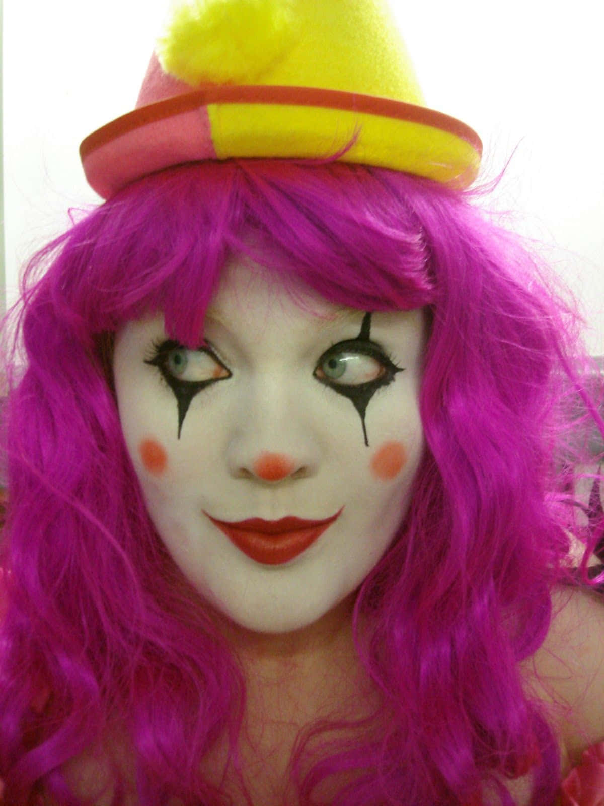 可愛女孩搞笑小丑圖片 壁紙 — 一個擁有鮮豔粉紅色頭髮和小丑妝容的人，在純色背景前戴著一頂色彩繽紛的帽子。