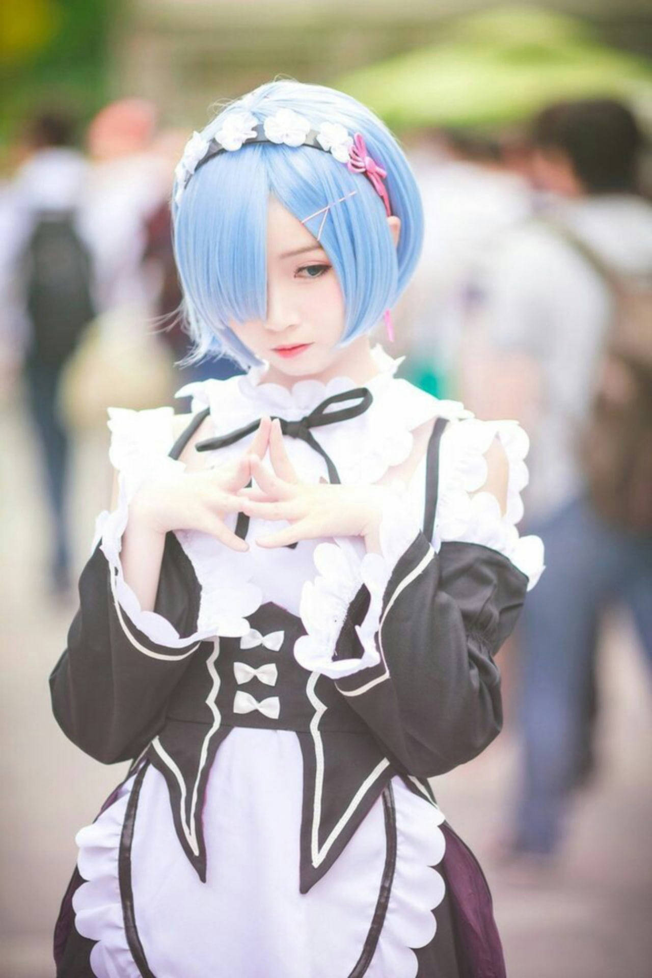 可愛女孩雷姆Cosplay 壁紙 — 一位藍髮女僕裝扮的cosplayer在模糊的戶外人群中用手比出心形。