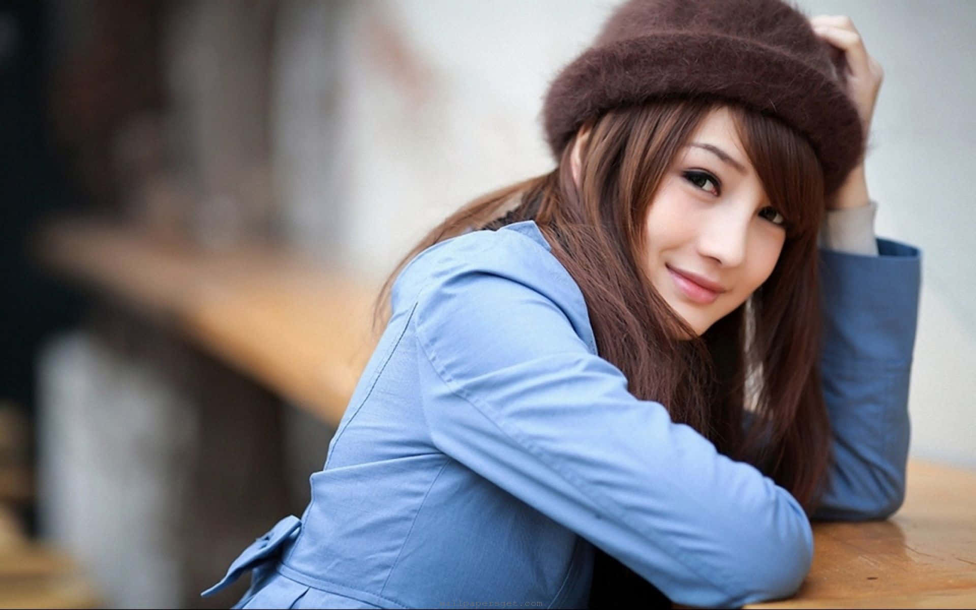 身穿藍色外套、戴著棕色帽子的年輕女性靠在木桌上微笑。